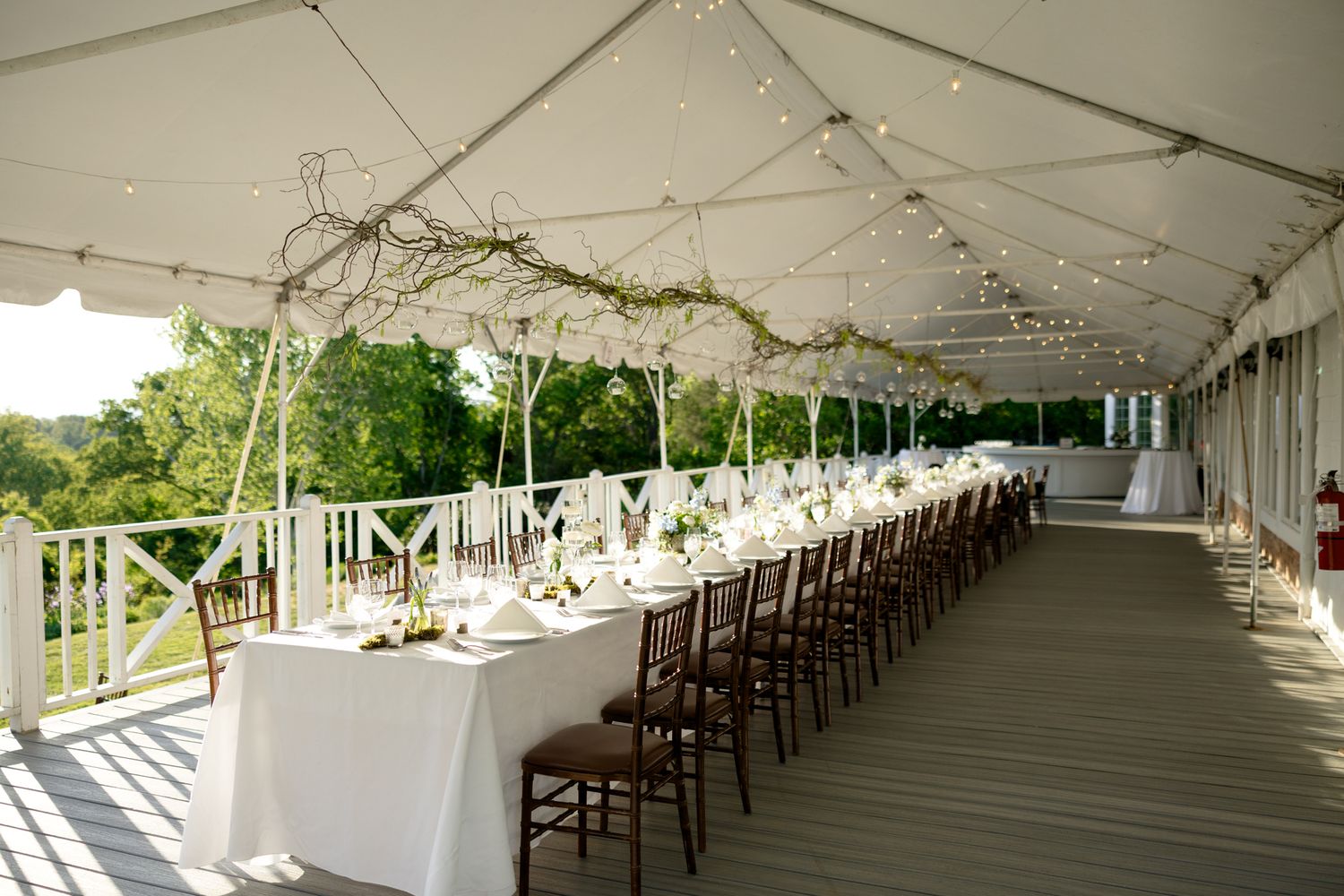 An elegant outdoor rehearsal dinner setup with long white tables under a tent with string lights and floral decor.