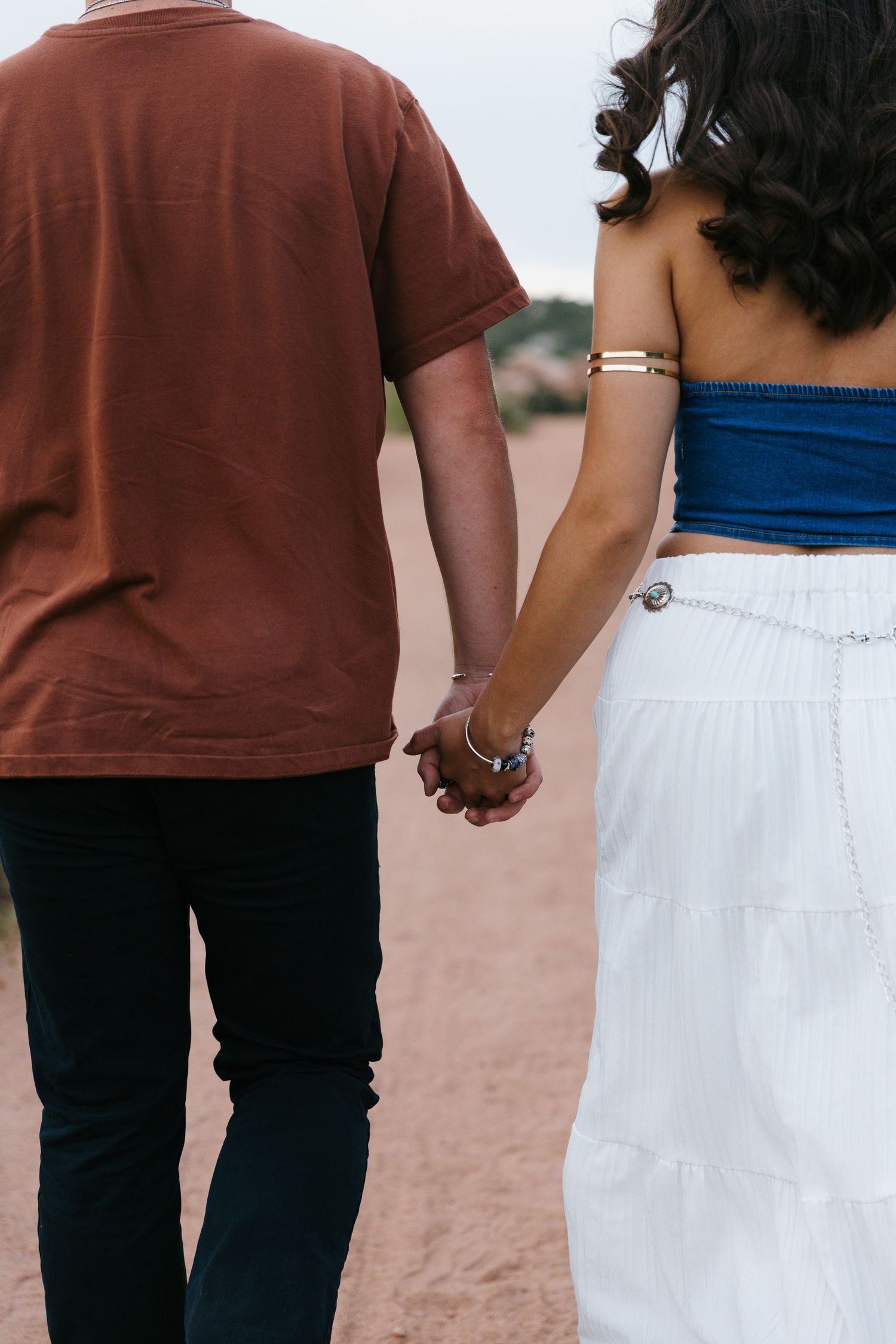 Couple holding hands from behind, woman in blue crop top and white skirt on sandy path.