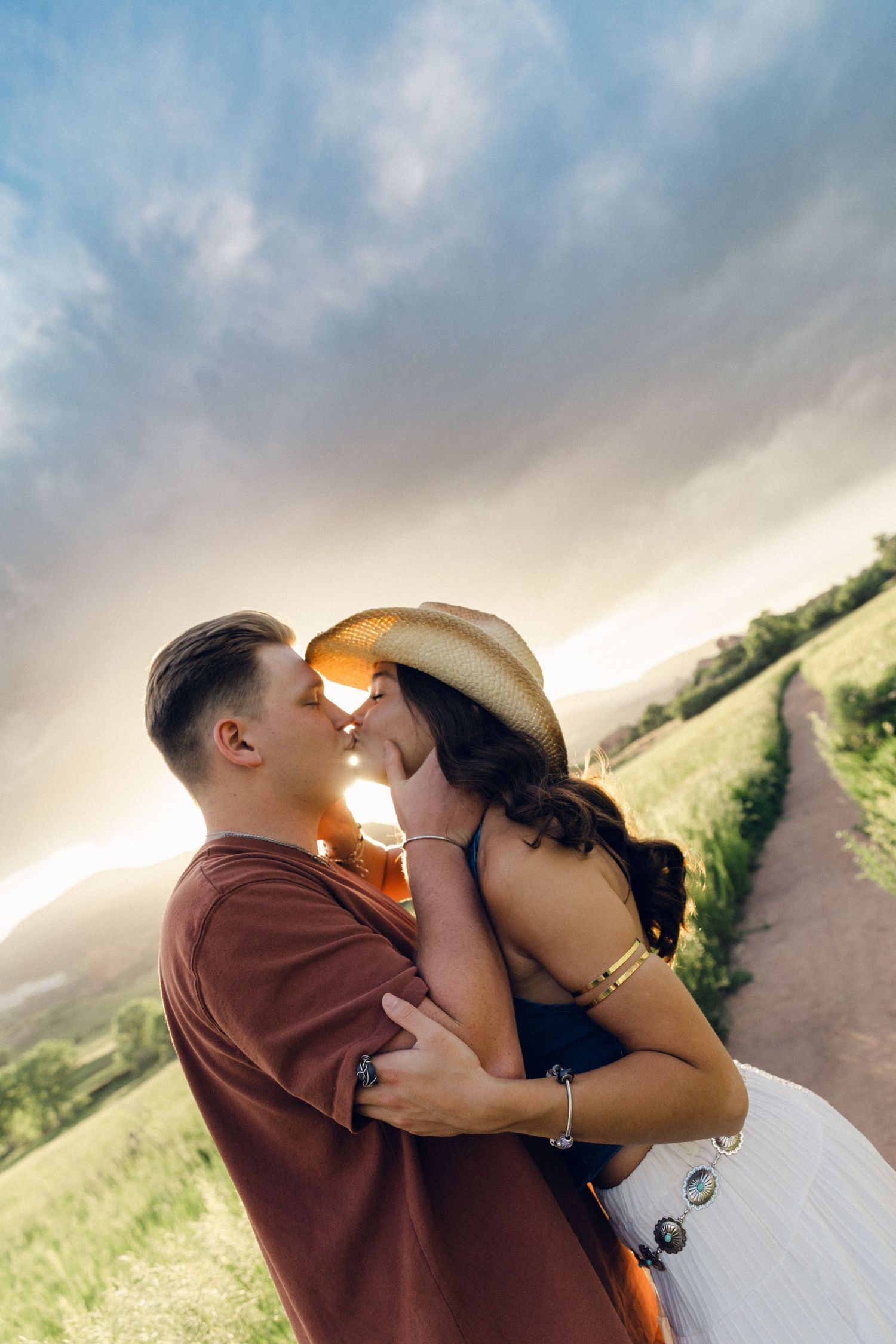 Romantic couple kissing under dramatic cloudy sky at sunset in a scenic rural landscape.