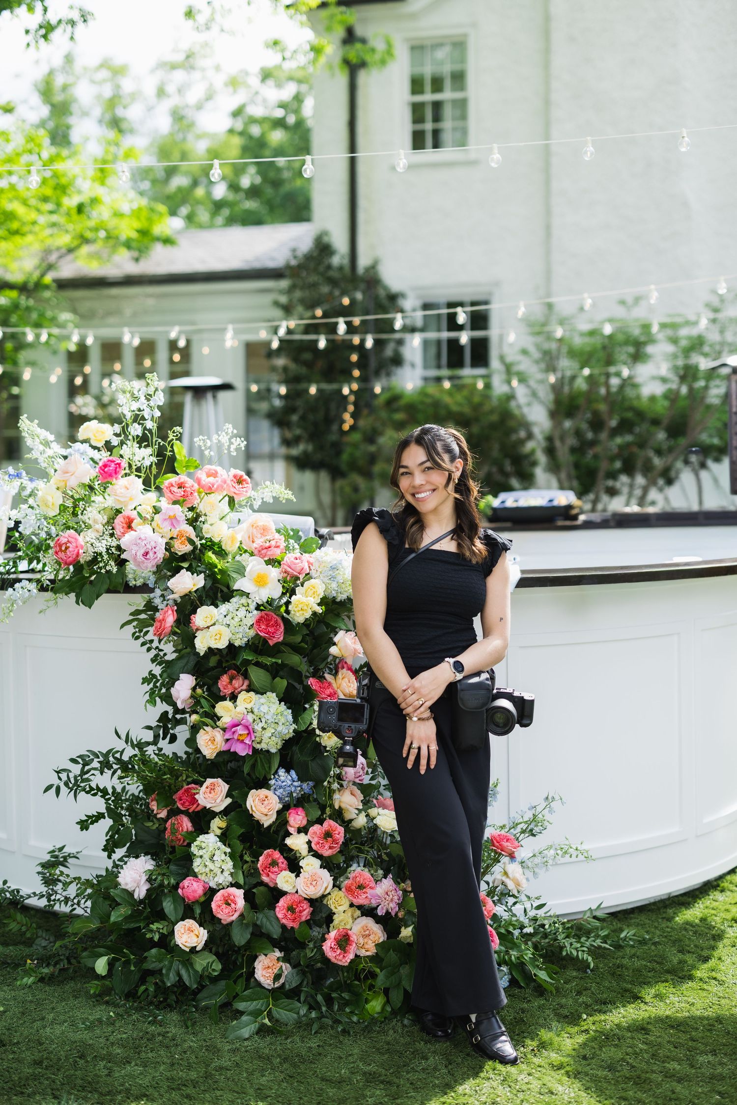 Woman in black jumpsuit holding camera poses beside colorful floral arrangement at elegant outdoor wedding venue.