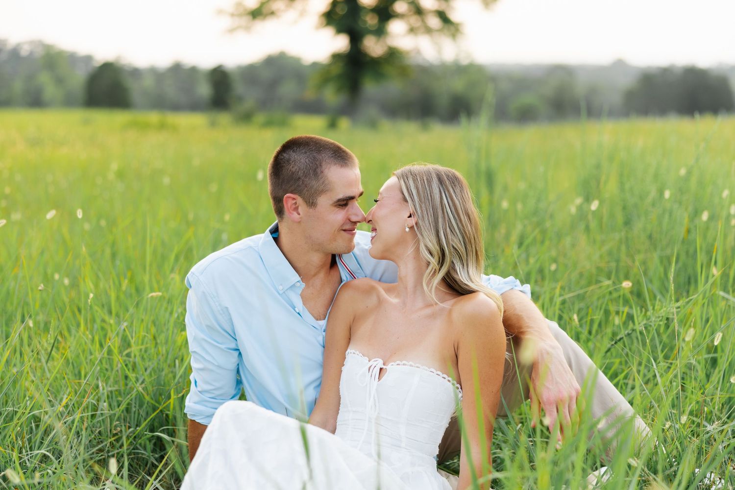 A couple sits intimately in a lush green field, sharing a romantic moment while dressed in casual white and blue attire.