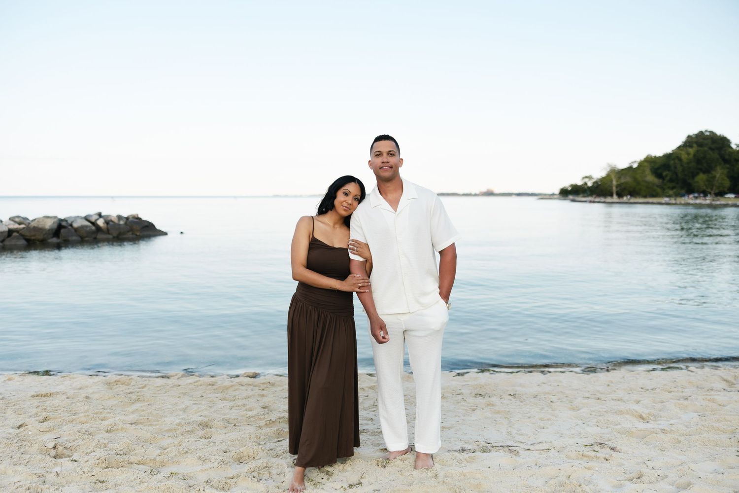 Couple posing on a sandy beach by calm blue water, woman in brown maxi dress, man in white outfit.