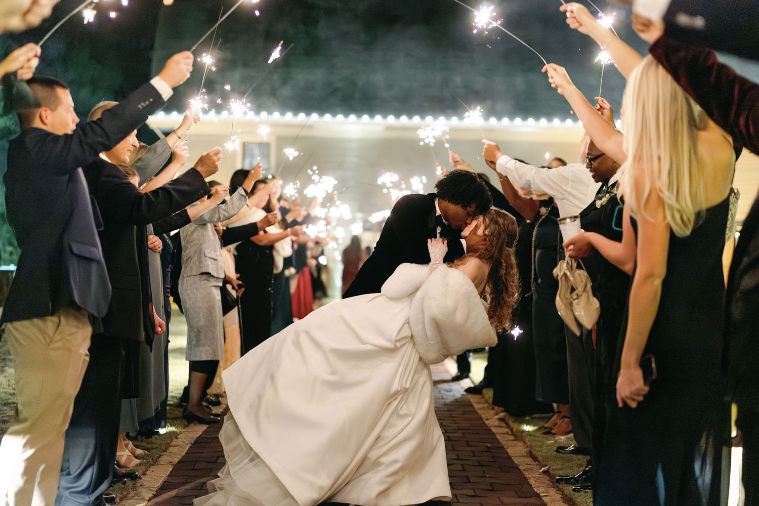 Newlyweds share a dramatic dip kiss surrounded by guests holding sparklers at their wedding exit.