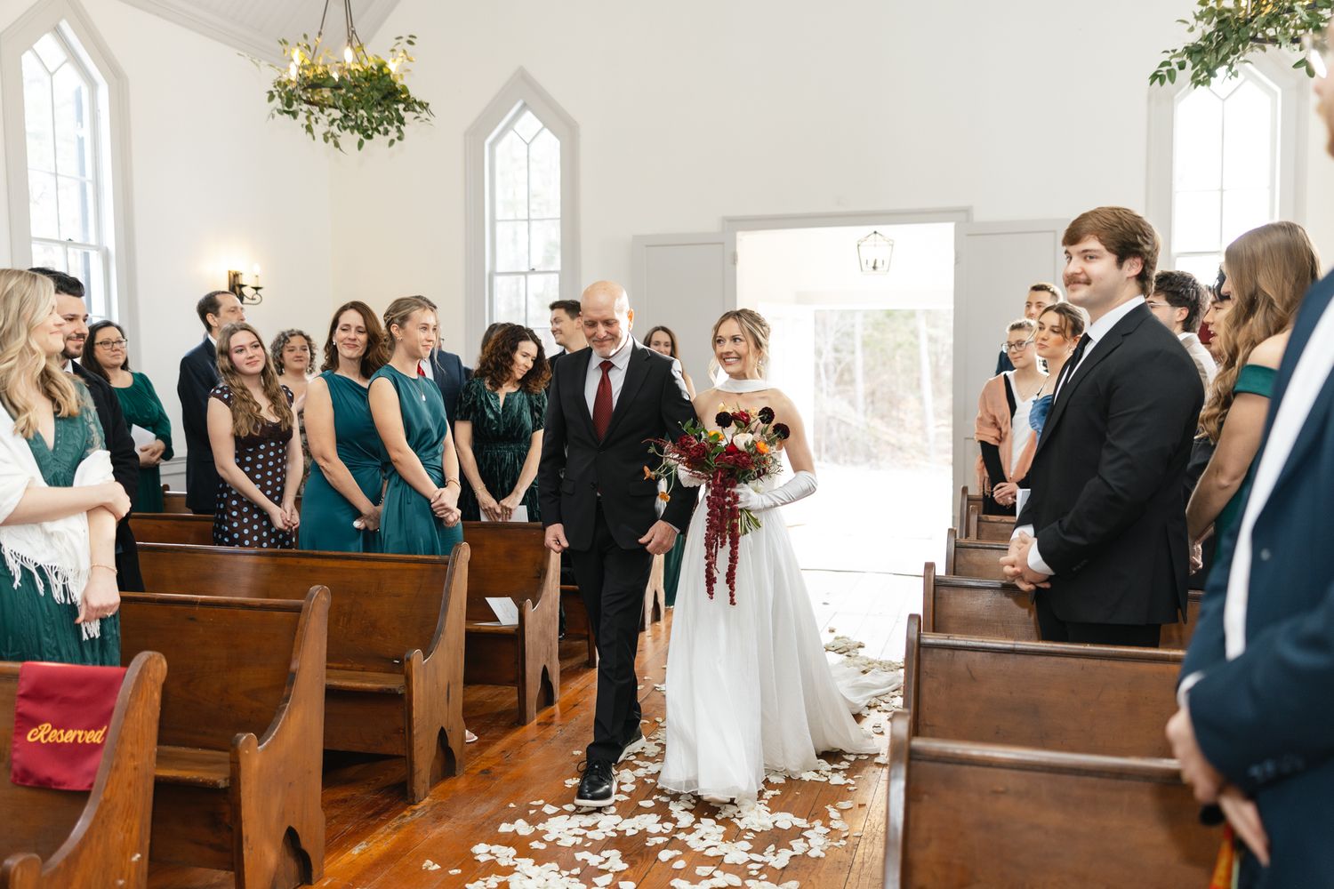 Bride walks down flower petal aisle with father as guests in green and burgundy attire look on.