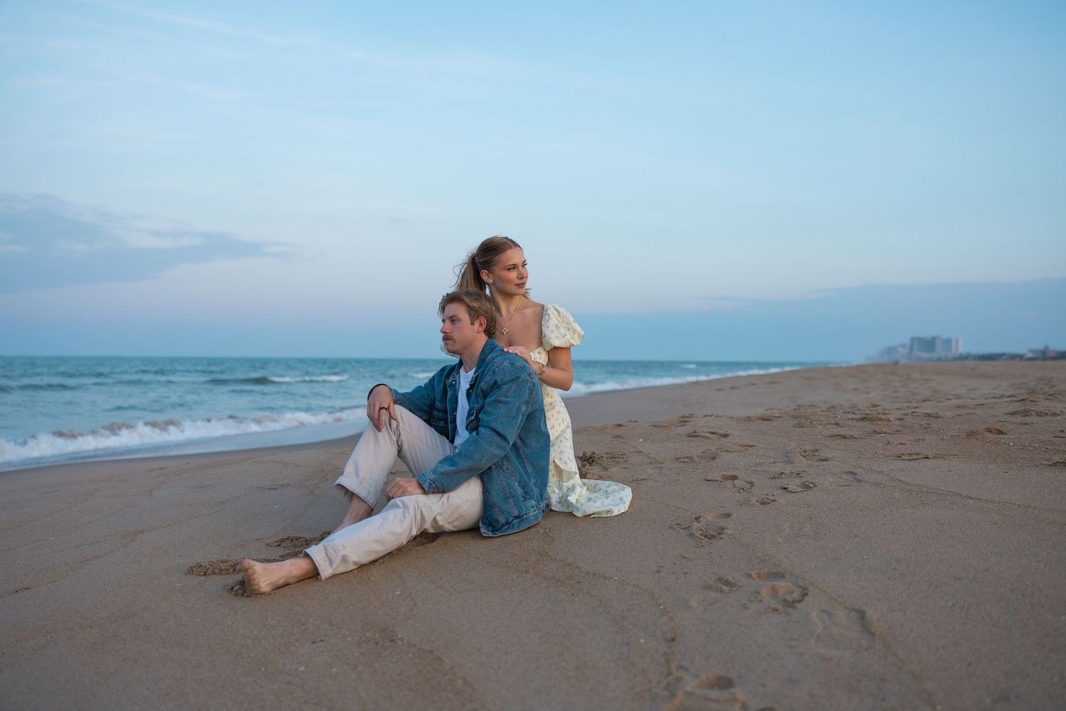 Couple sitting back-to-back on sandy beach at sunset with ocean waves behind them.