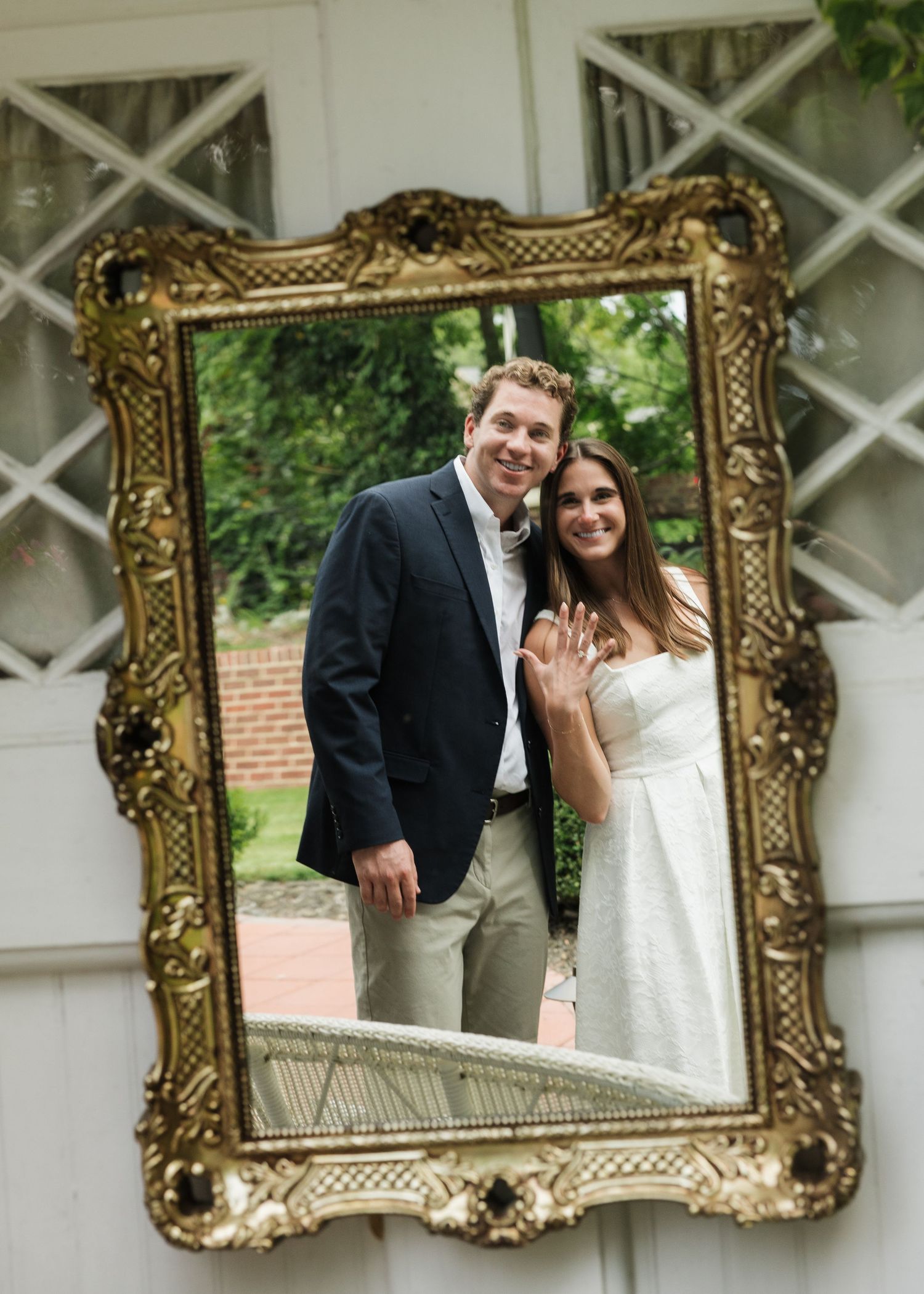 Couple smiling in ornate gold mirror, woman showing engagement ring, wearing white dress and navy blazer outdoors.