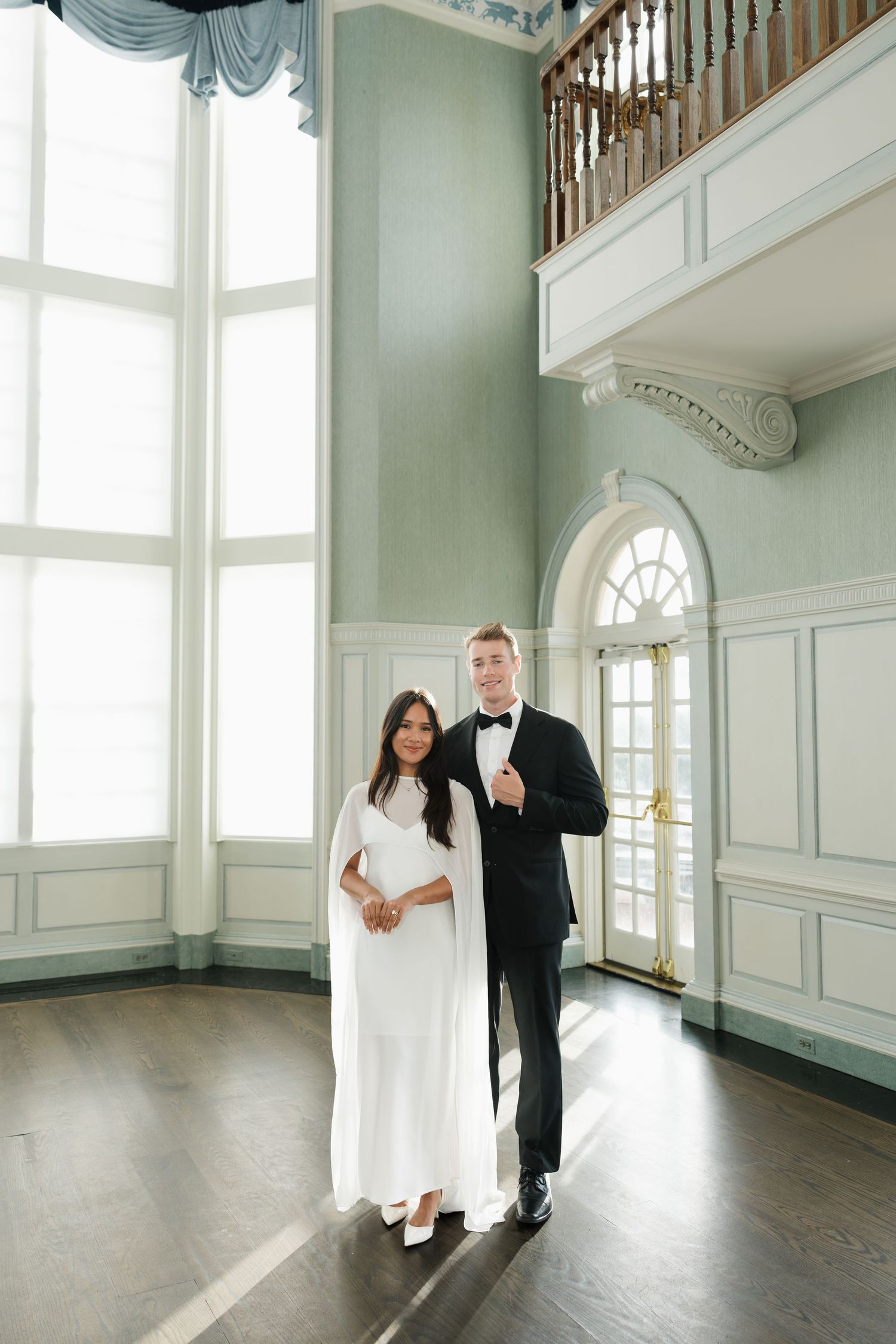 Elegant couple in wedding attire posing in a grand ballroom with high ceilings and large windows.