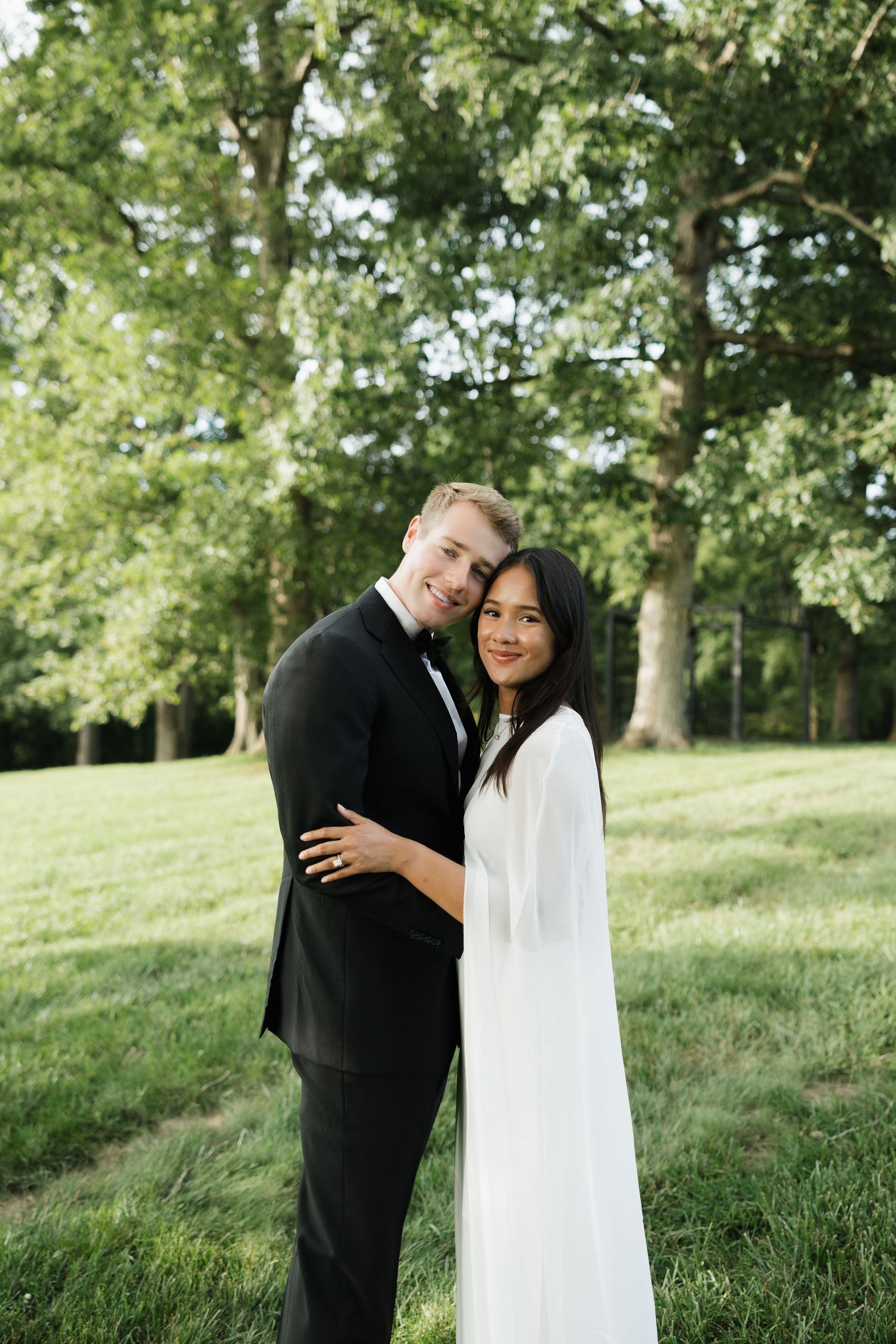 A couple in wedding attire embraces outdoors, groom in black tuxedo and bride in white gown, with lush green trees behind.