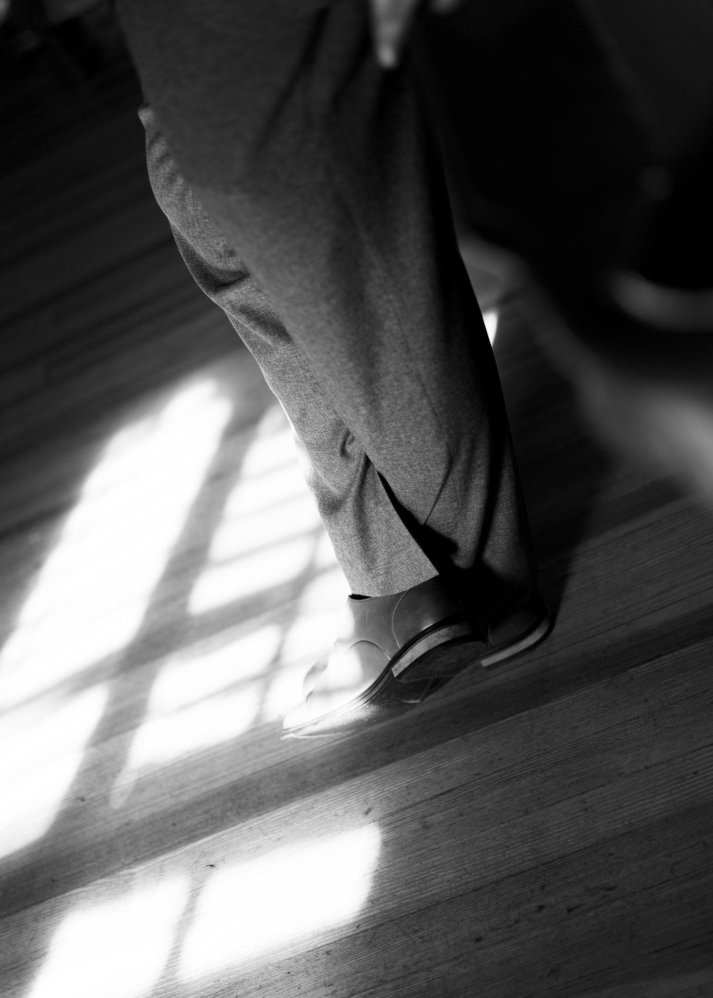 Black and white photo of person in grey trousers and dress shoes standing on sunlit hardwood floor.