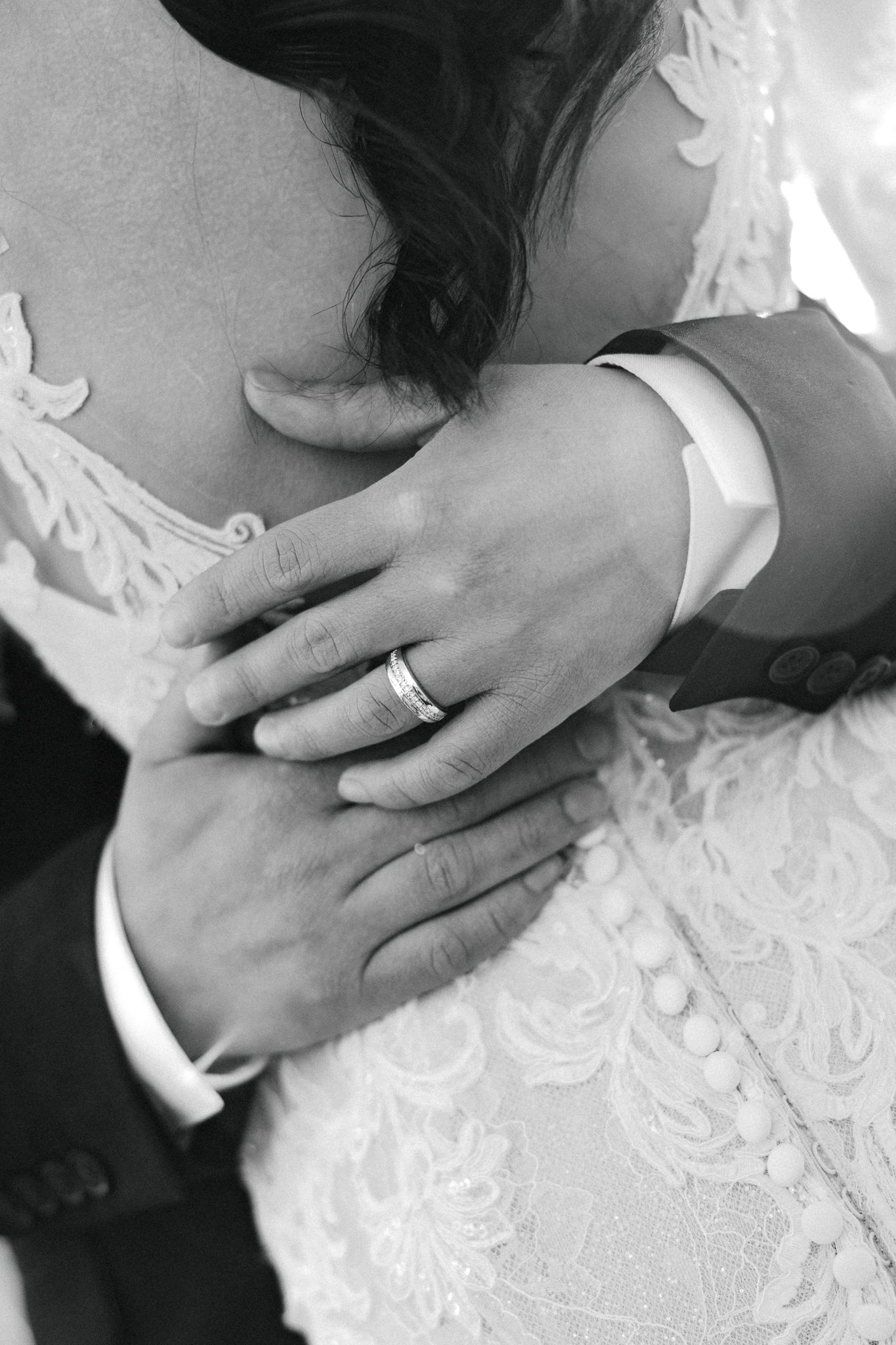 Black and white close-up of grooms hand with wedding band embracing brides lace gown.