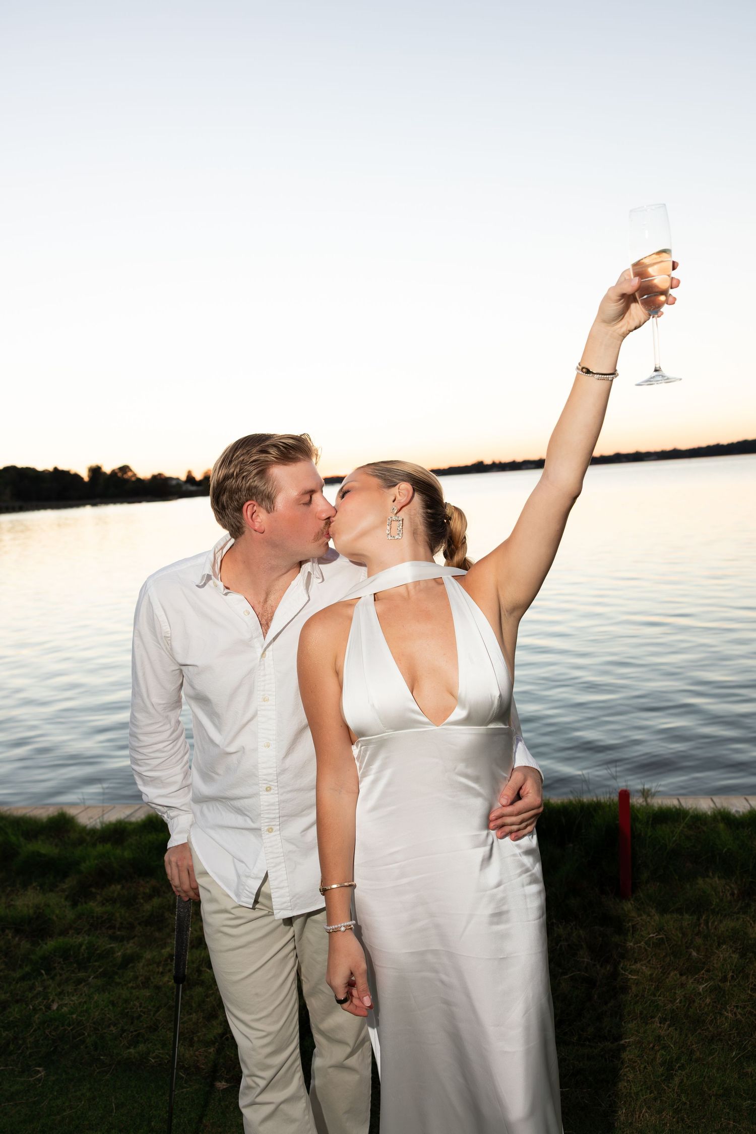 A couple kisses at sunset by a lake, woman in white dress raising a champagne glass in celebration.
