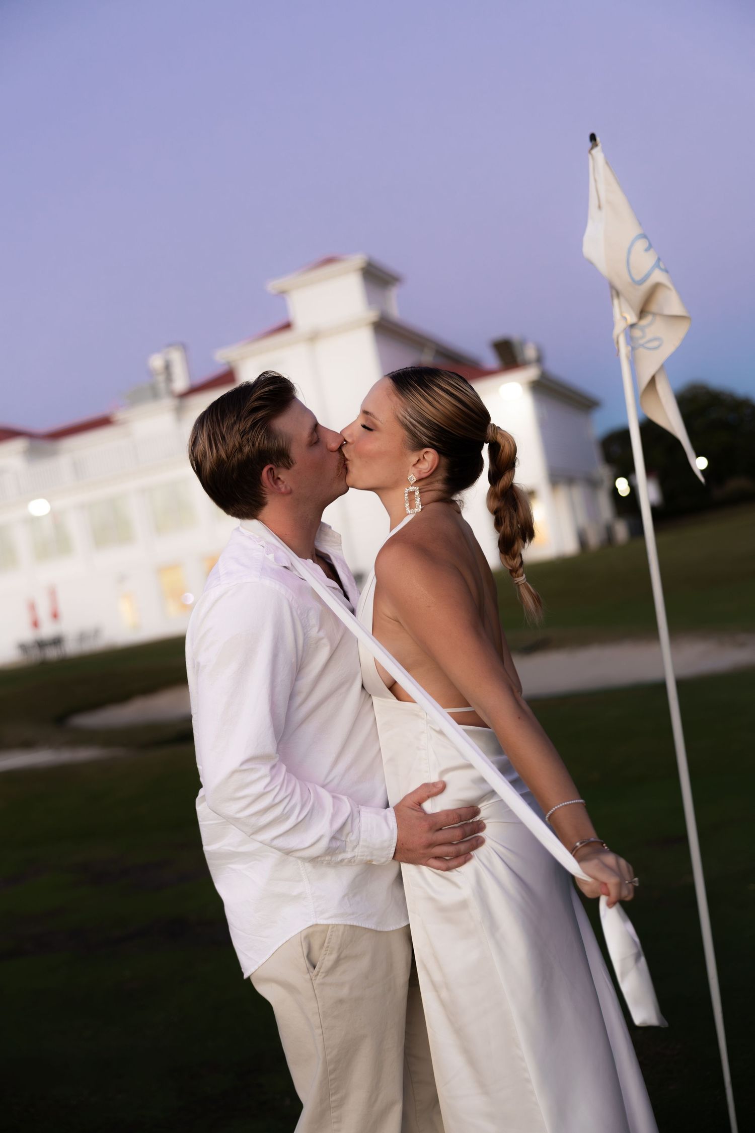 Couple in white attire sharing a romantic kiss on a golf course at dusk, with a elegant white building in the background.