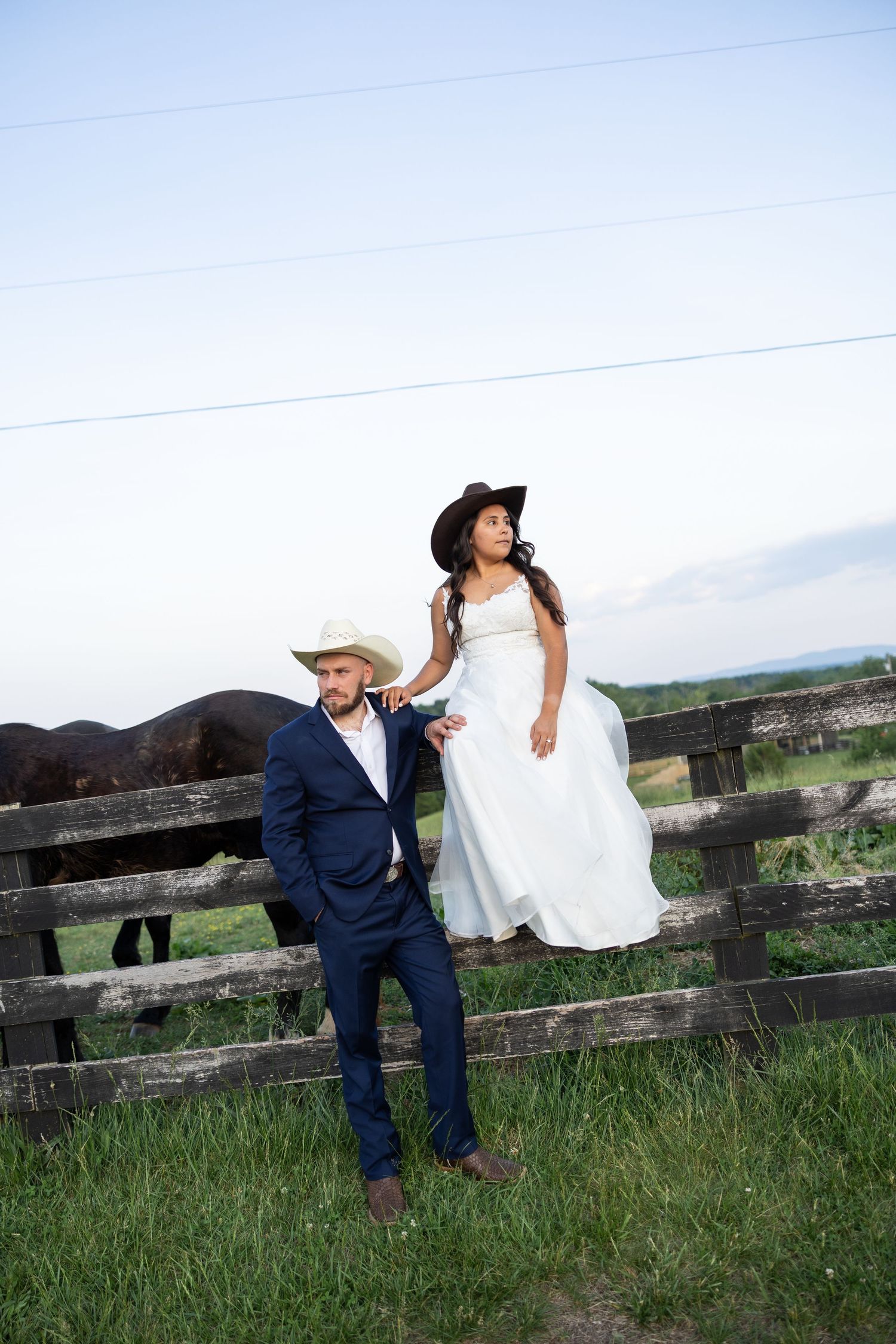 Bride and groom pose by wooden fence on farm, both wearing cowboy hats with cattle grazing behind them.