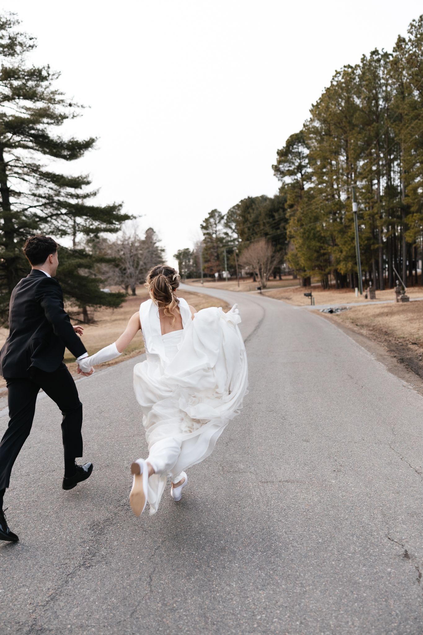 Bride and groom run hand-in-hand down a tree-lined road in wedding attire during outdoor photo session.