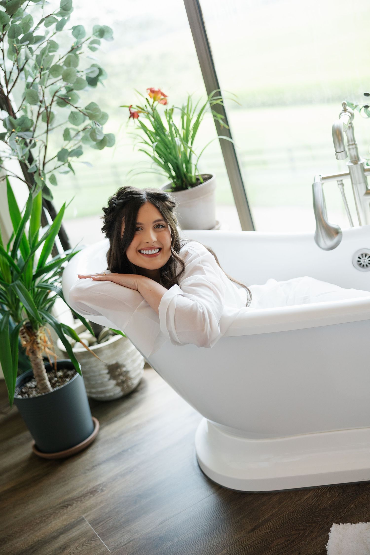 Smiling bride relaxing in a freestanding white bathtub surrounded by lush green plants in a modern bathroom.