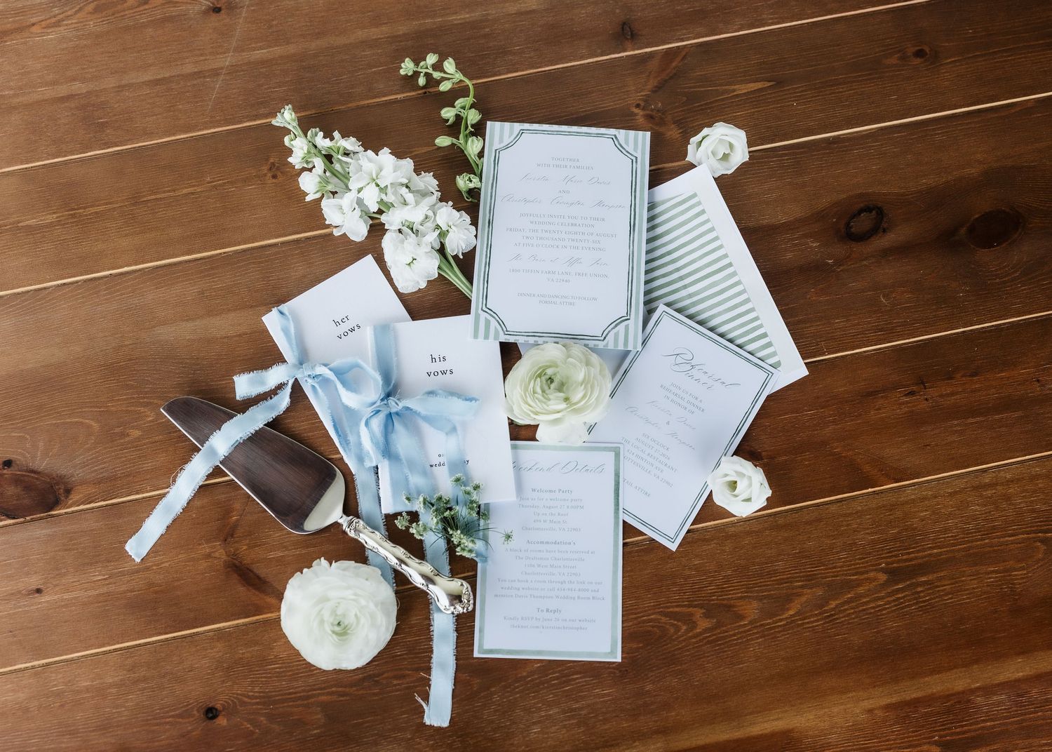 Elegant wedding stationery suite with blue accents, white florals, cake server, and ribbon on rustic wood floor.