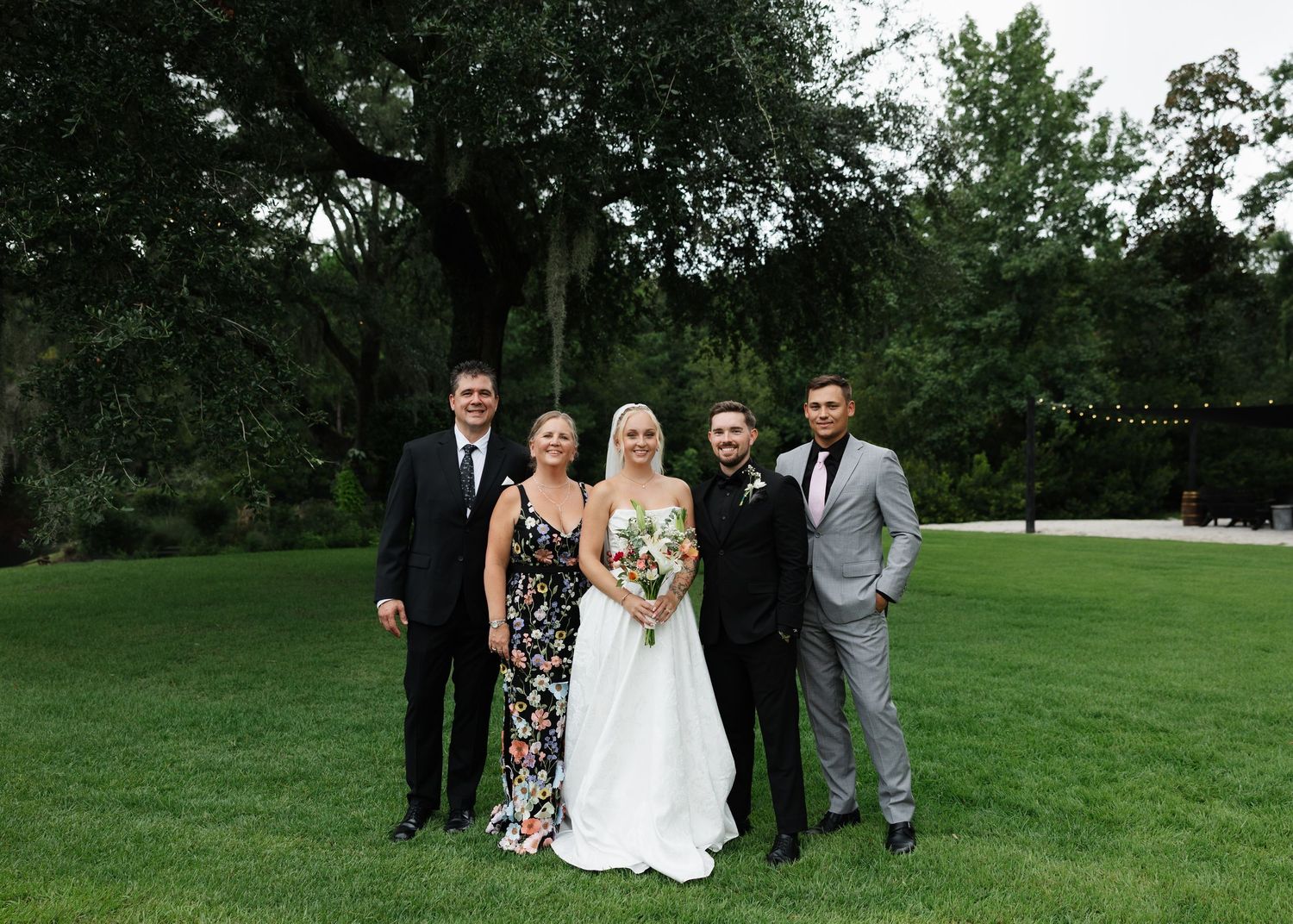 Wedding party poses outdoors on green lawn with large trees, bride in white gown holding bouquet with four guests.