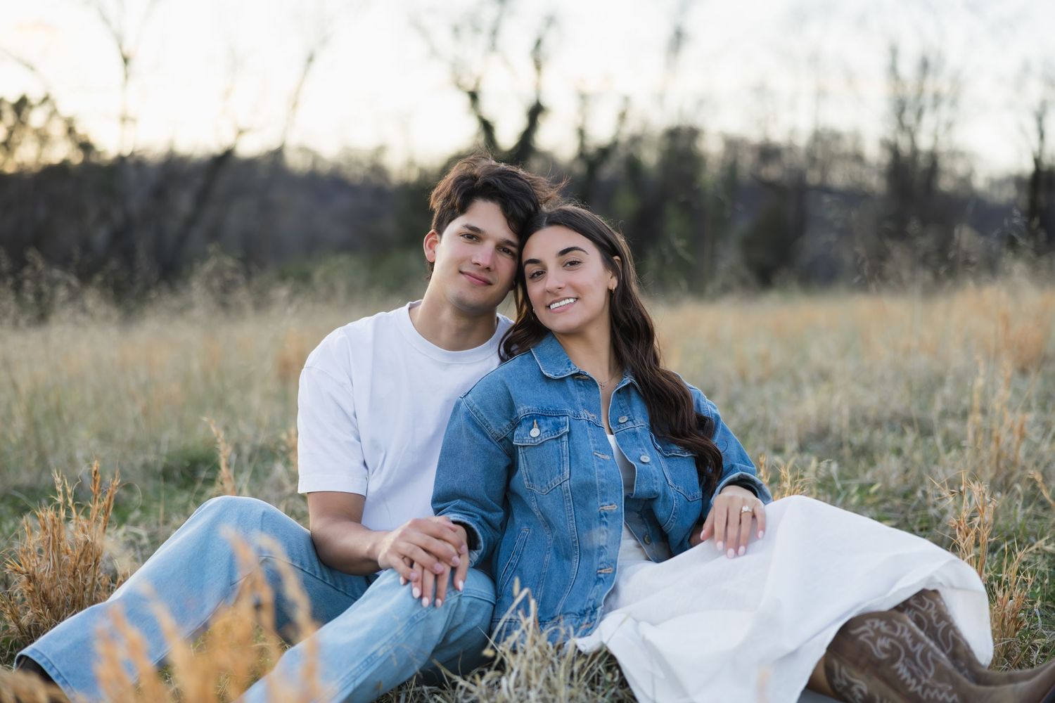 A close-up of a young couple sitting in a field, smiling warmly while holding hands in soft golden light.