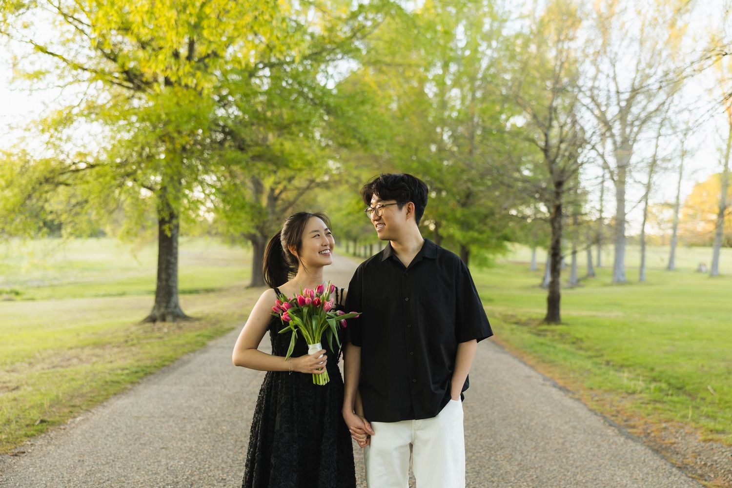 A smiling couple walks hand-in-hand on a park path, the woman in black holding red tulips amid lush green trees.