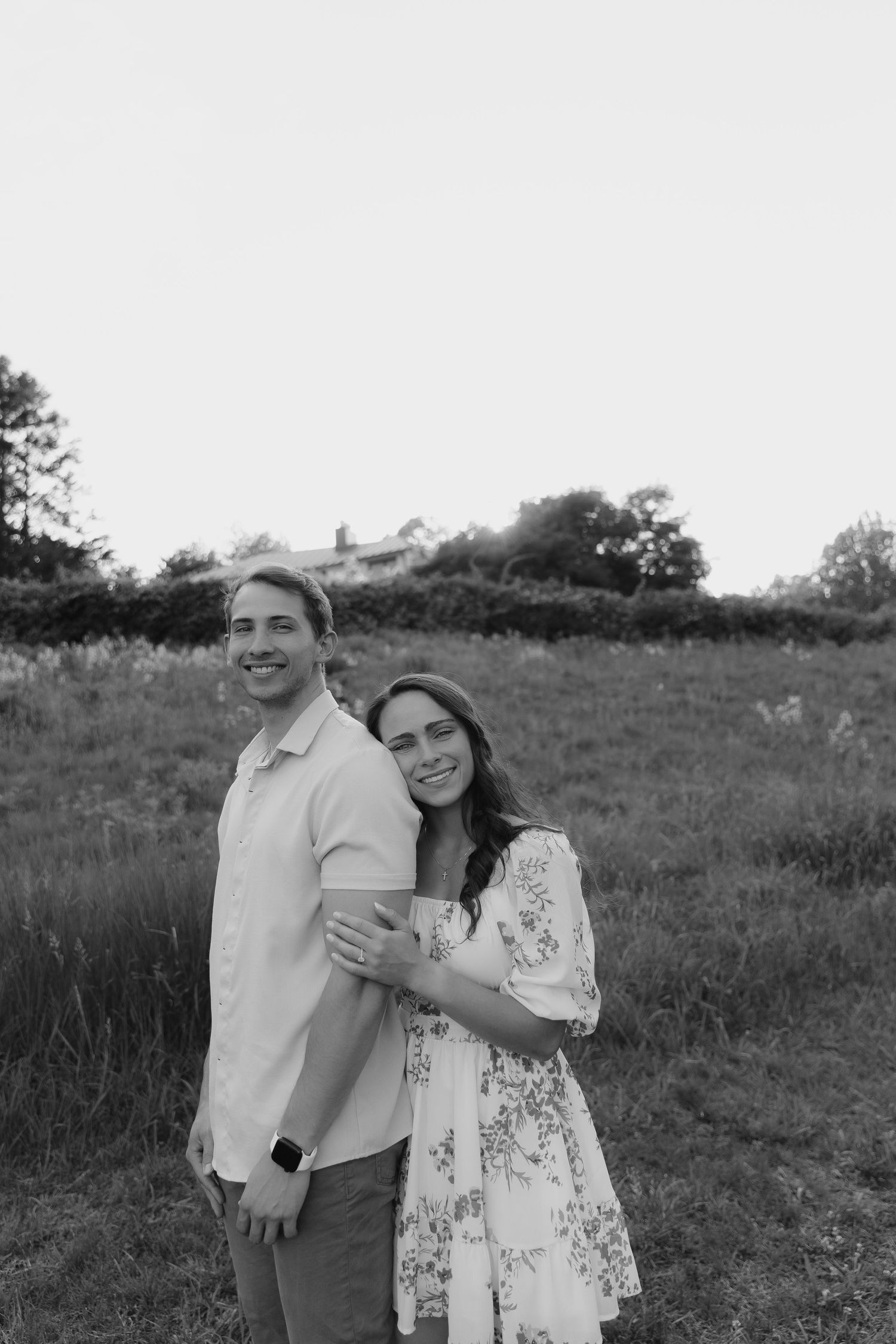 Black and white photo of a smiling couple embracing in a meadow at golden hour.