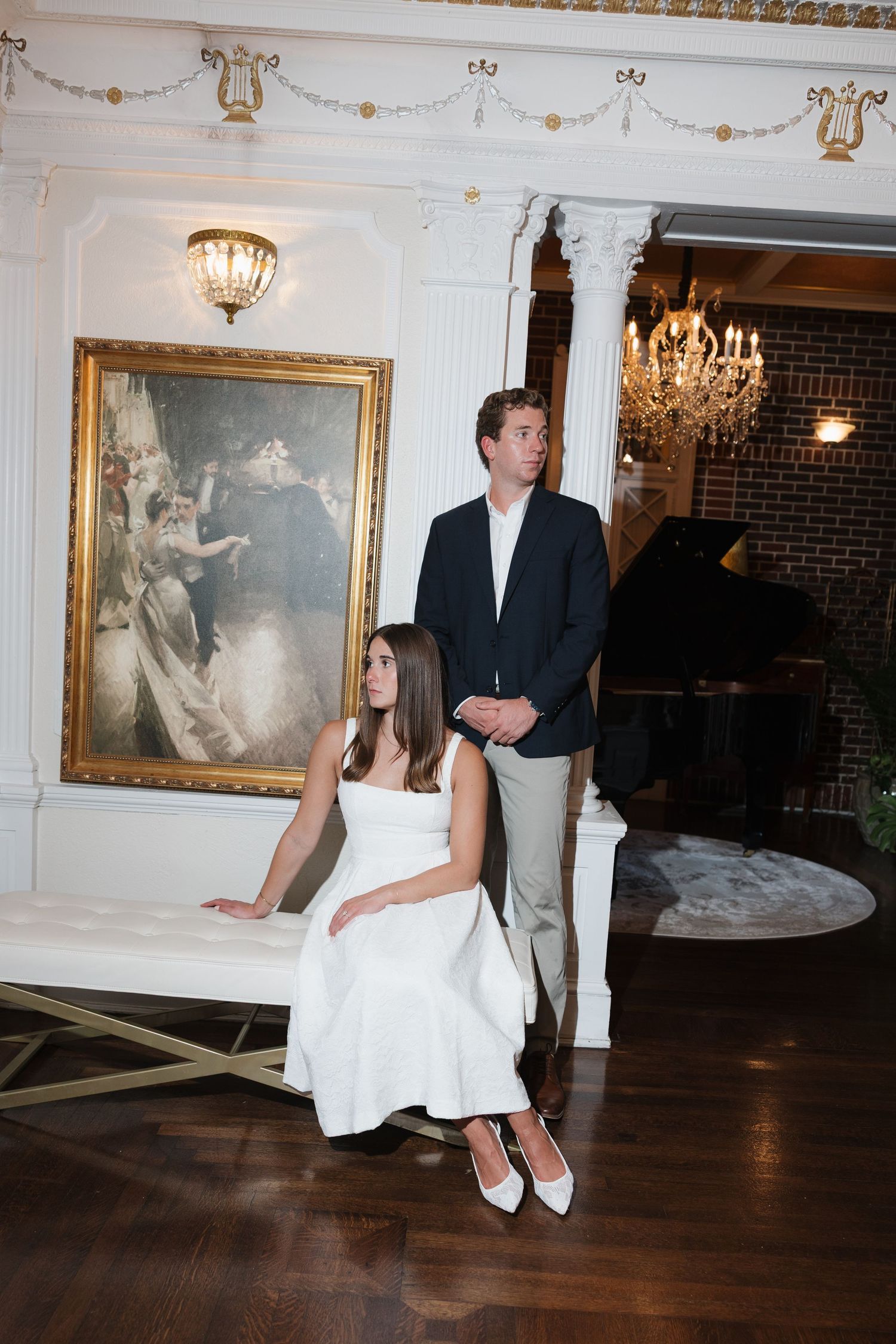 Elegant couple in formal attire posing in ornate room with chandelier, large painting, and grand piano.