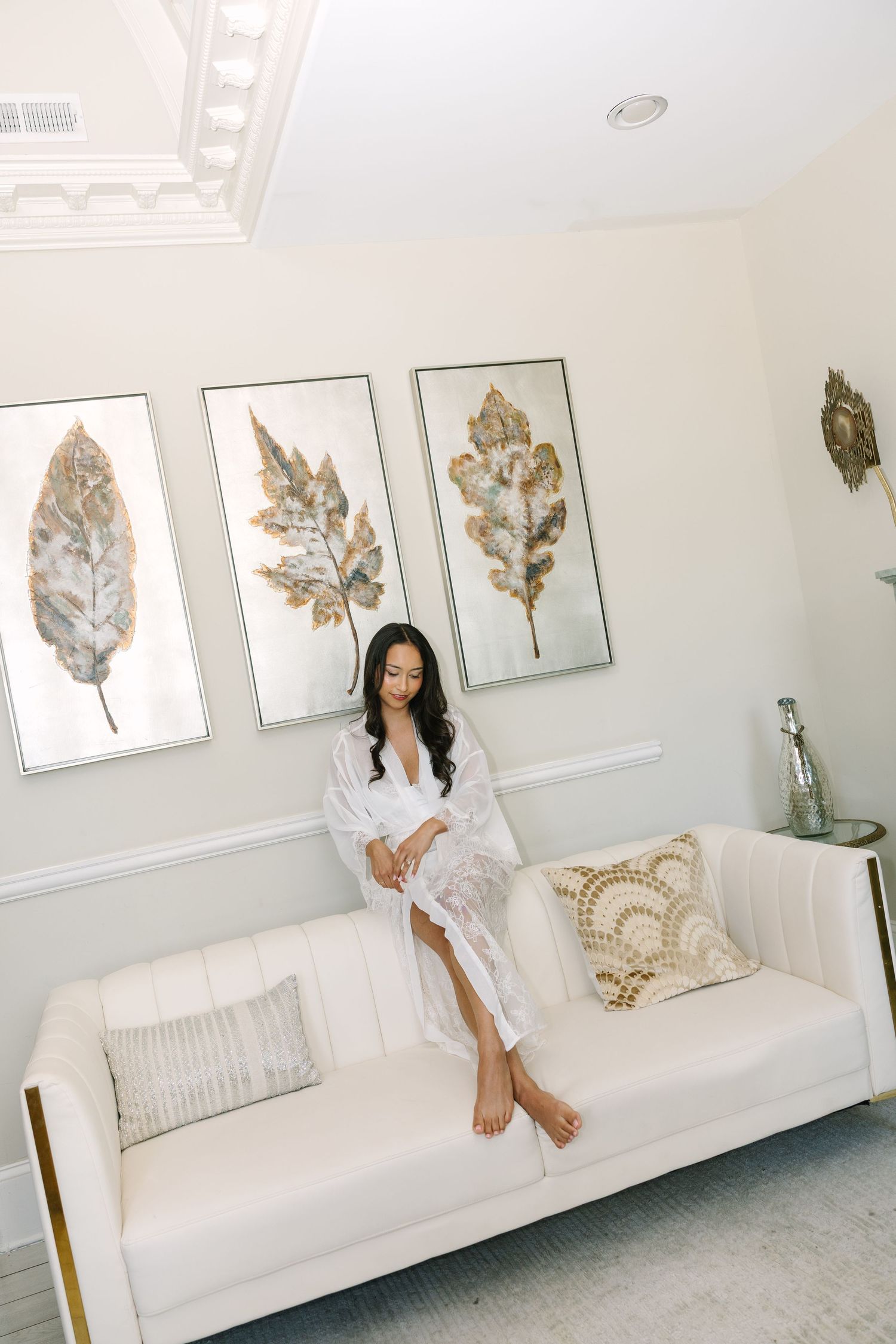 Woman in white robe sitting on cream sofa with gold leaf wall art in elegant bright room.