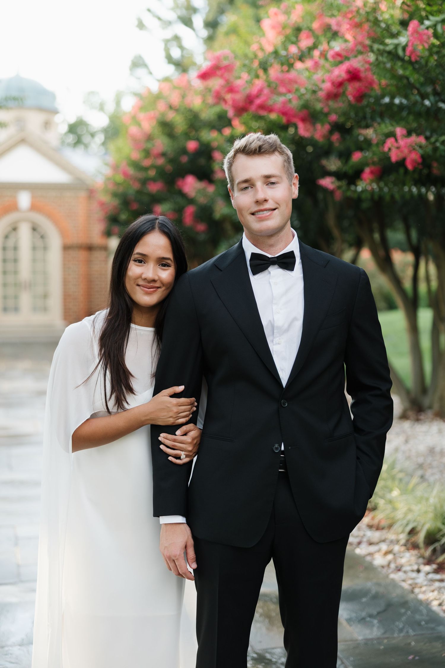 Elegant couple posing outdoors, woman in white dress and man in black tuxedo with bow tie, pink flowering trees behind.