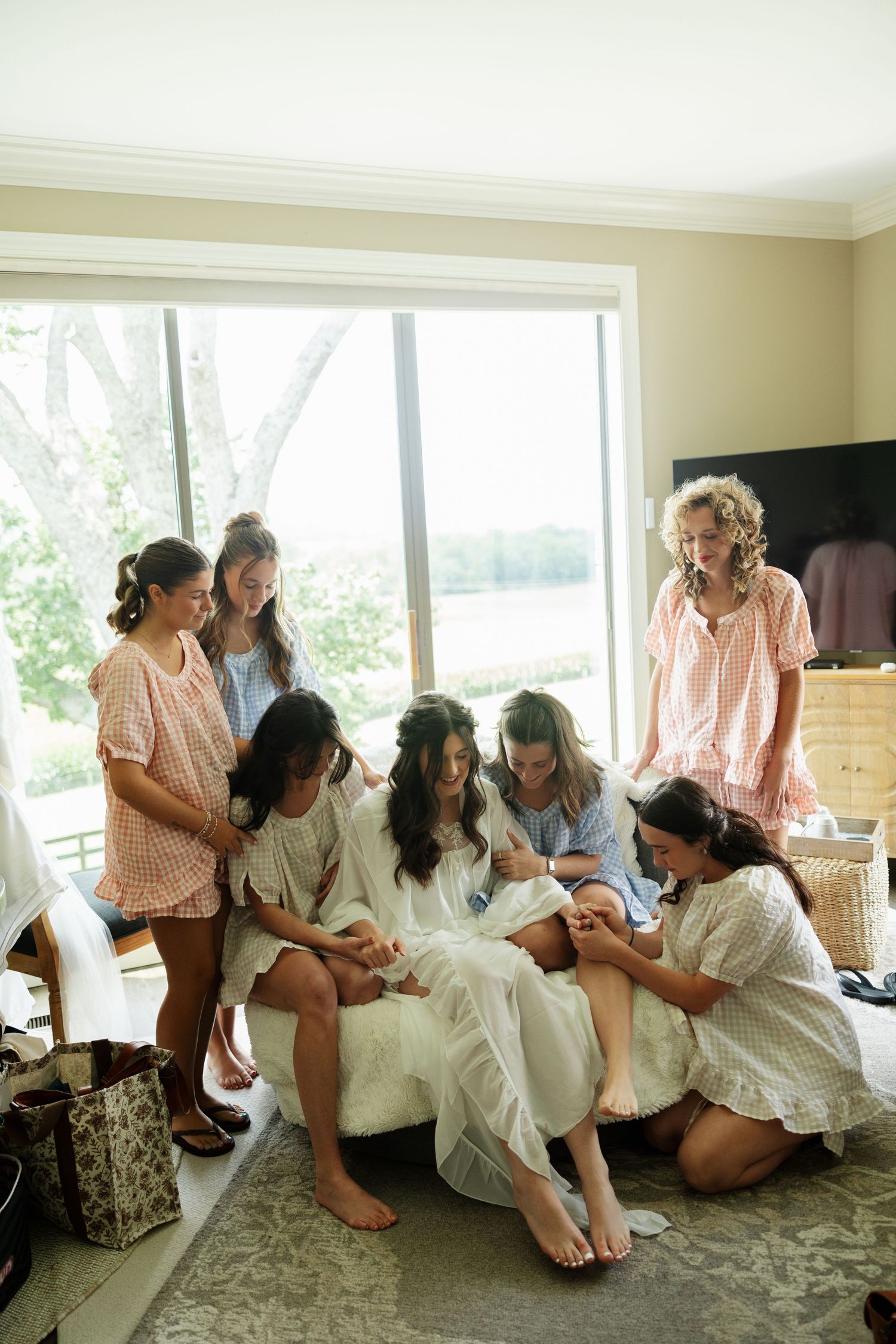 Bride and bridesmaids in robes gather together on a couch during wedding morning preparations indoors.