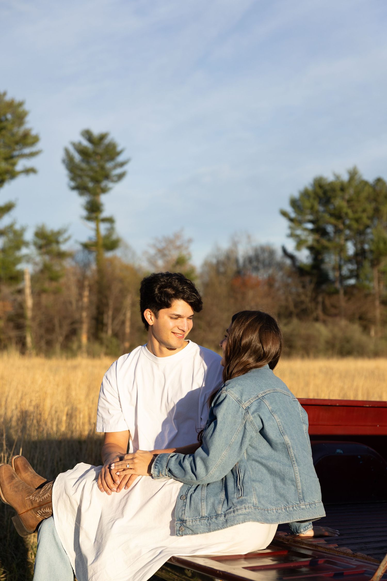 Young couple sitting on red truck tailgate, smiling at each other in a sunny field.