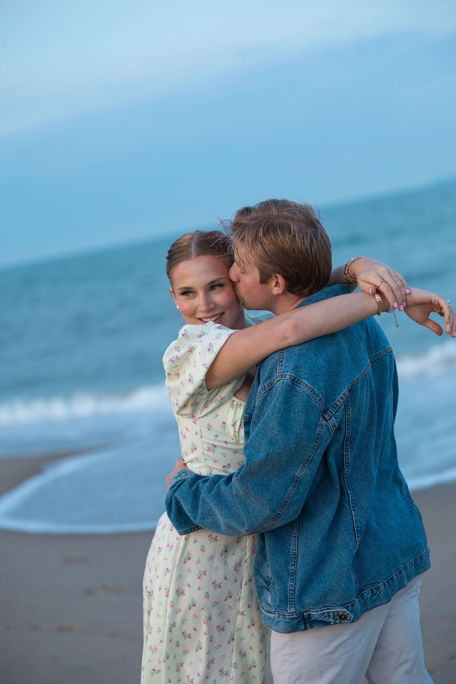 Man kissing womans cheek while embracing on sandy beach at twilight near ocean waves.