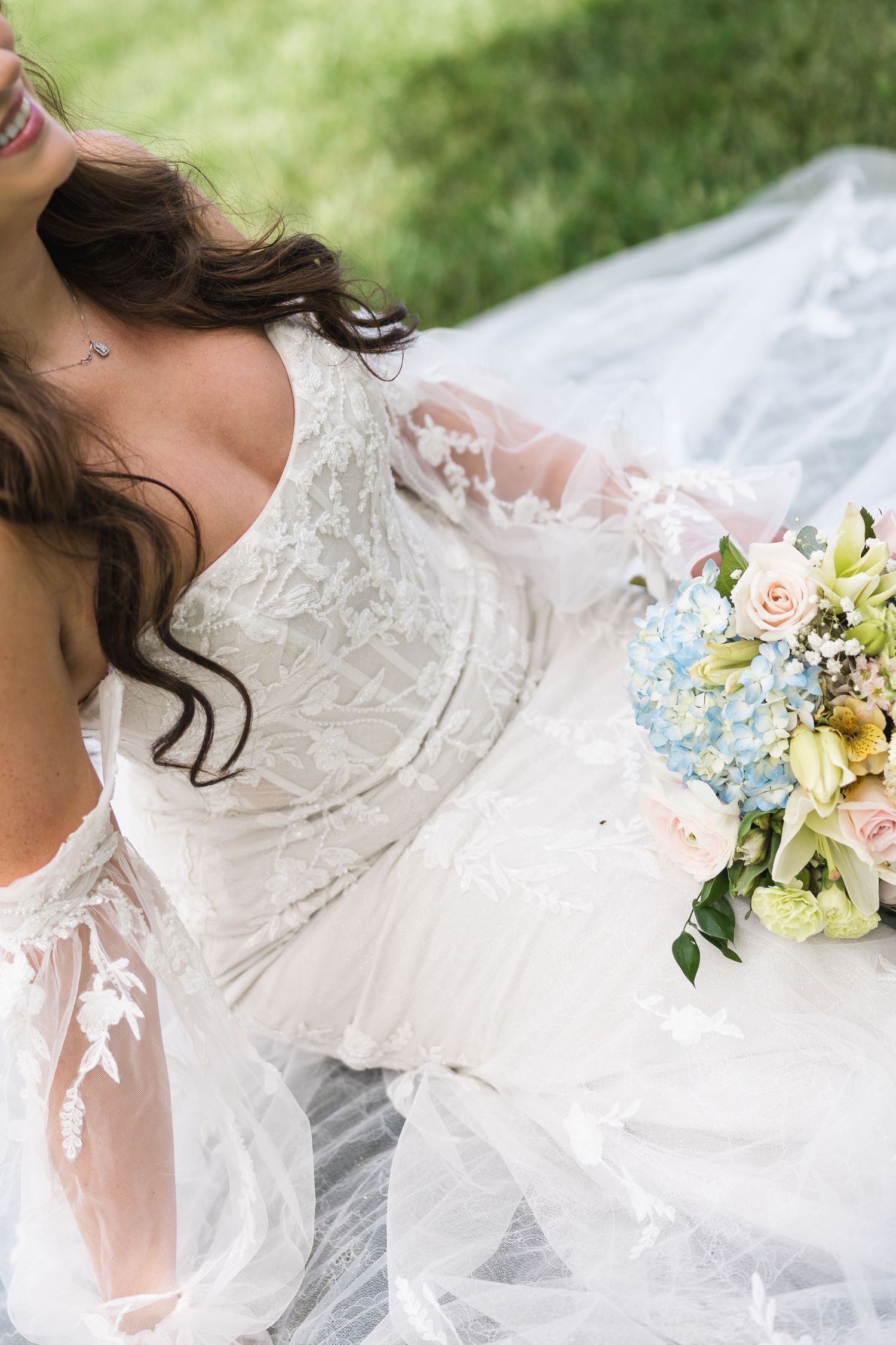 Bride in lace wedding gown sitting outdoors holding colorful bouquet of blue hydrangeas and pink roses.