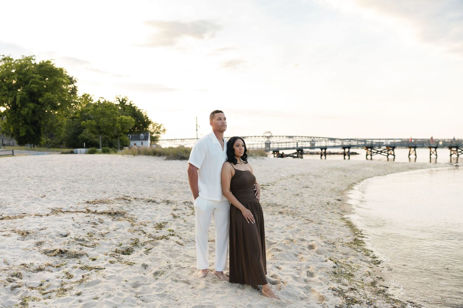 Pregnant couple posing on white sand beach at sunset, man in white outfit, woman in brown maxi dress, marina in background.