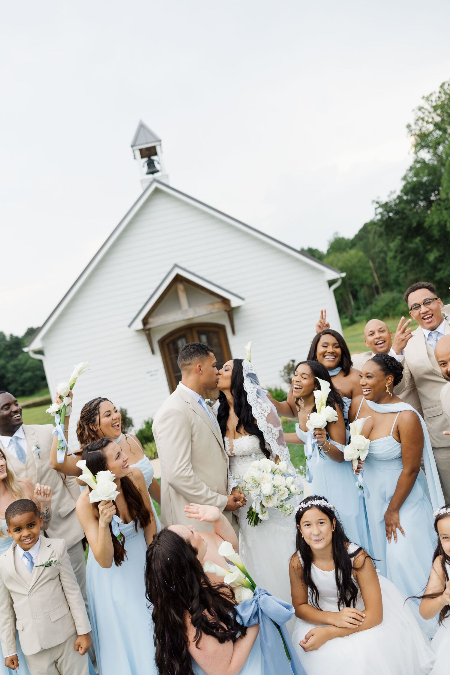 Wedding party in light blue dresses and tan suits celebrating outside a white chapel, bride and groom kissing in center.