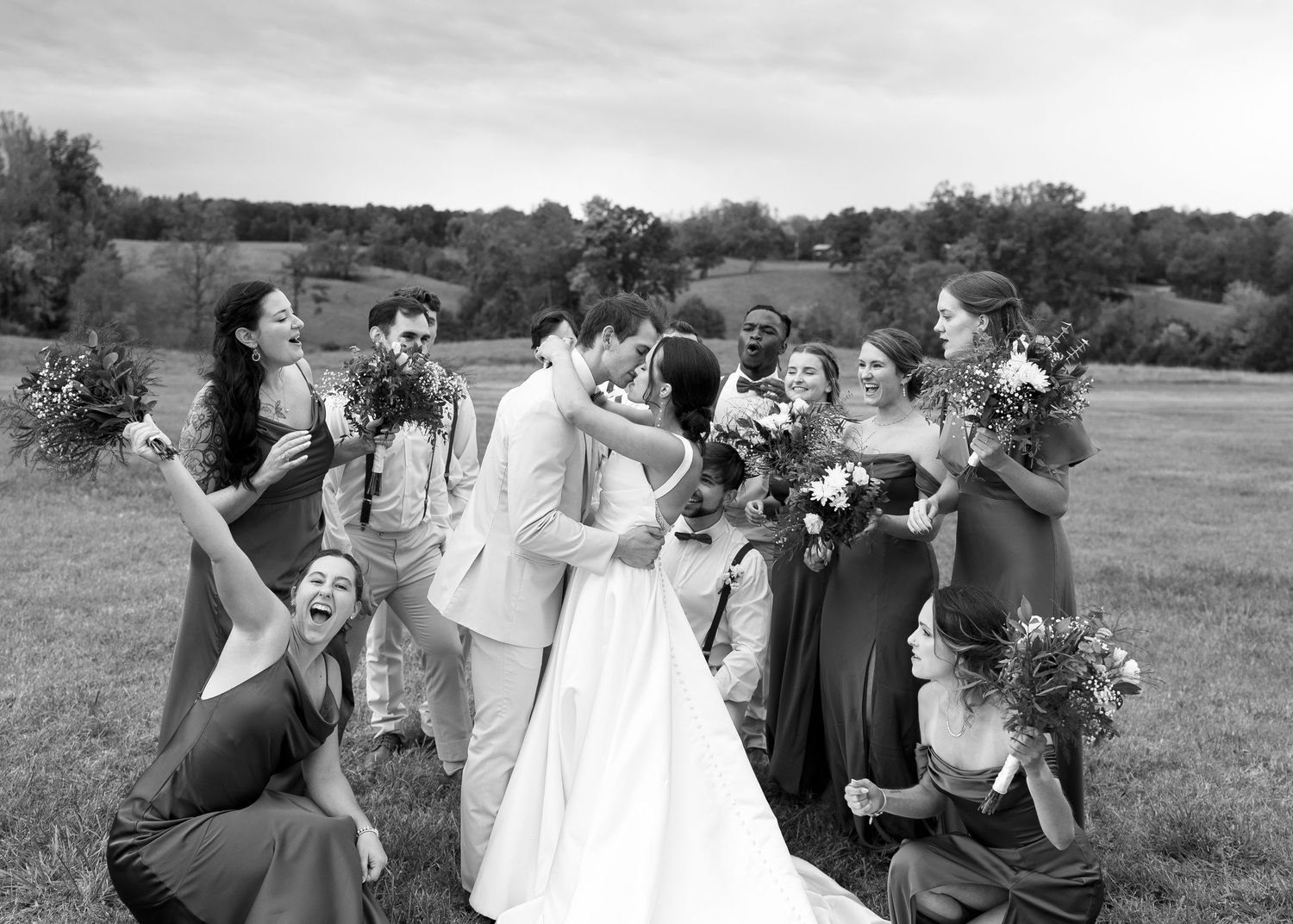 Wedding party celebrating as bride and groom kiss in a lush green field, bridesmaids in sage gowns holding bouquets.