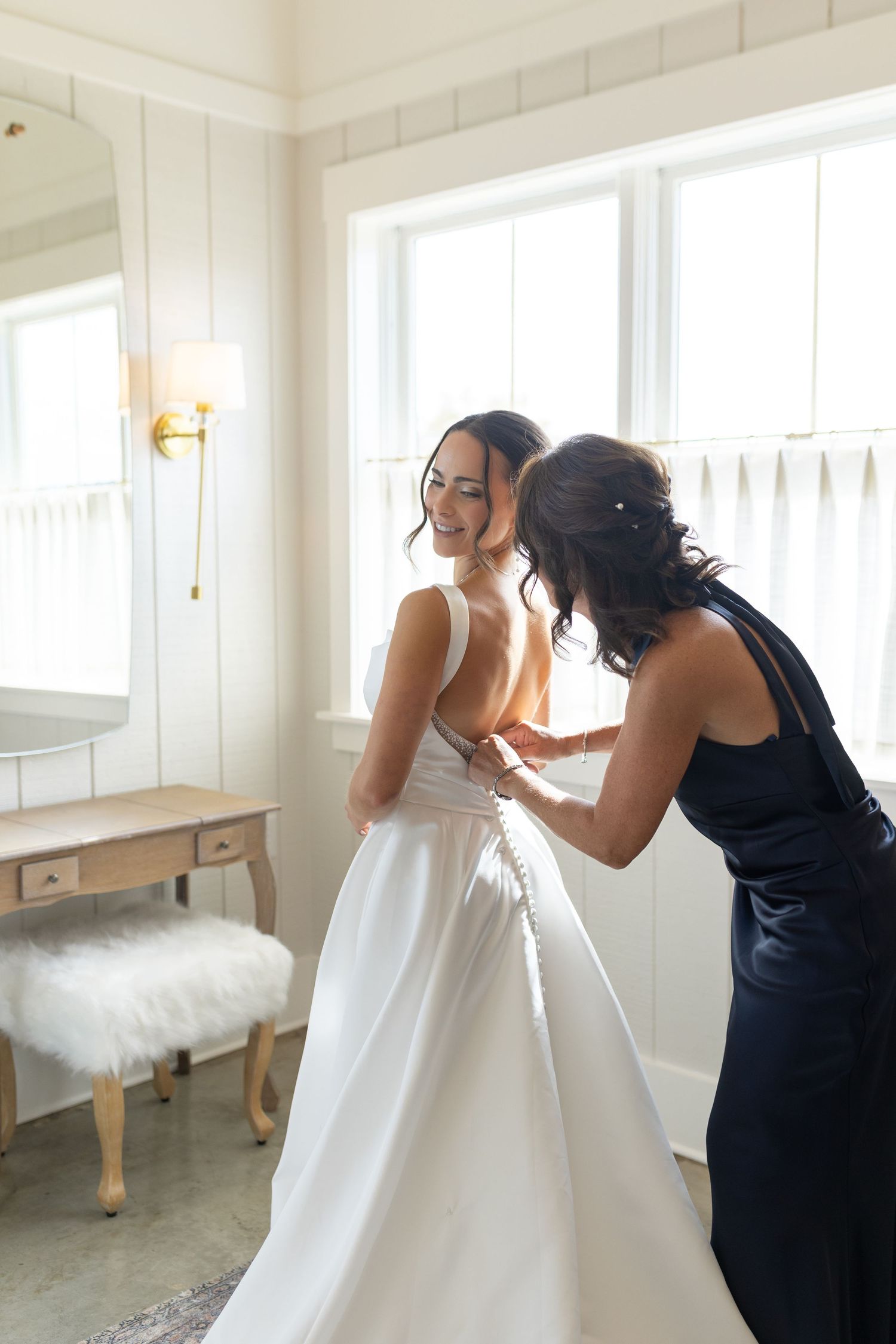 Bridesmaid fastening the back of the brides white gown in a sunlit dressing room before the ceremony.