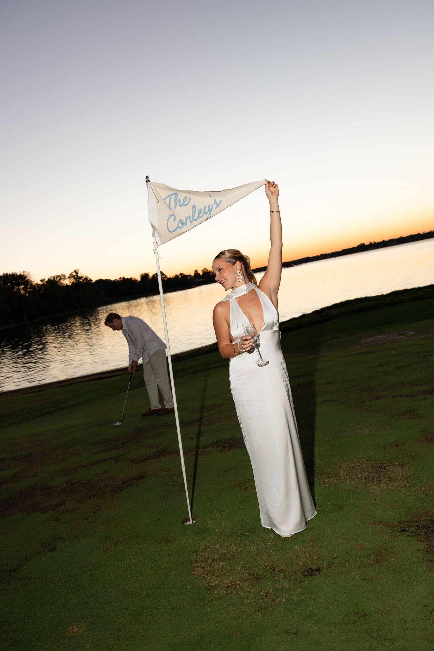 Bride in white gown holds golf flag reading The Conleys at sunset on a waterfront golf course.