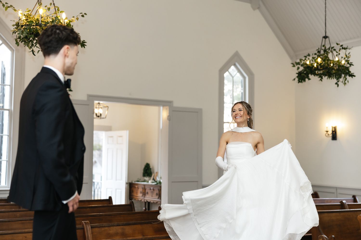 Joyful bride in white strapless gown twirls dress for groom inside elegant white chapel interior.