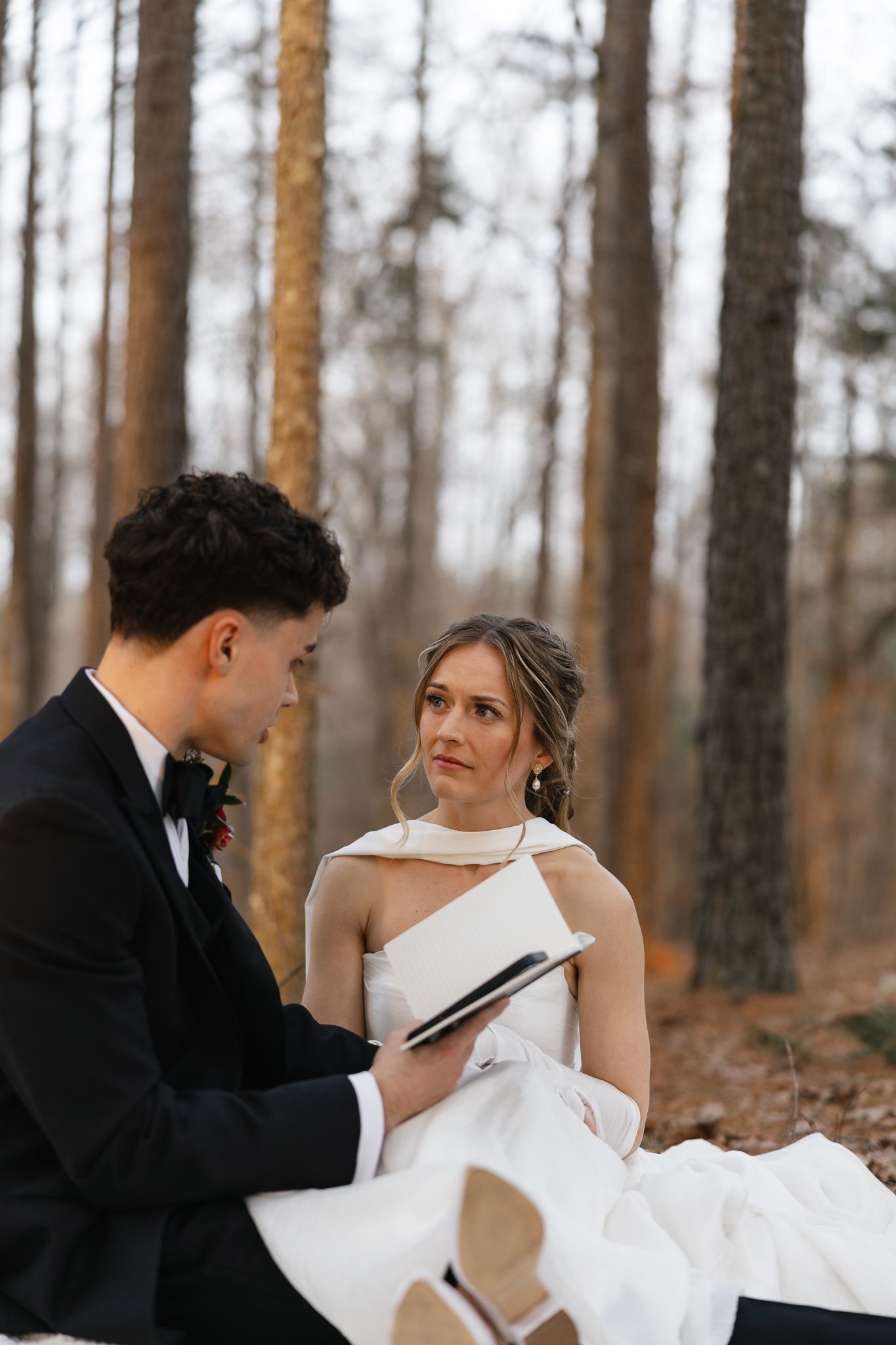 Bride and groom exchange vows from a black vow book while seated in an autumn pine forest.