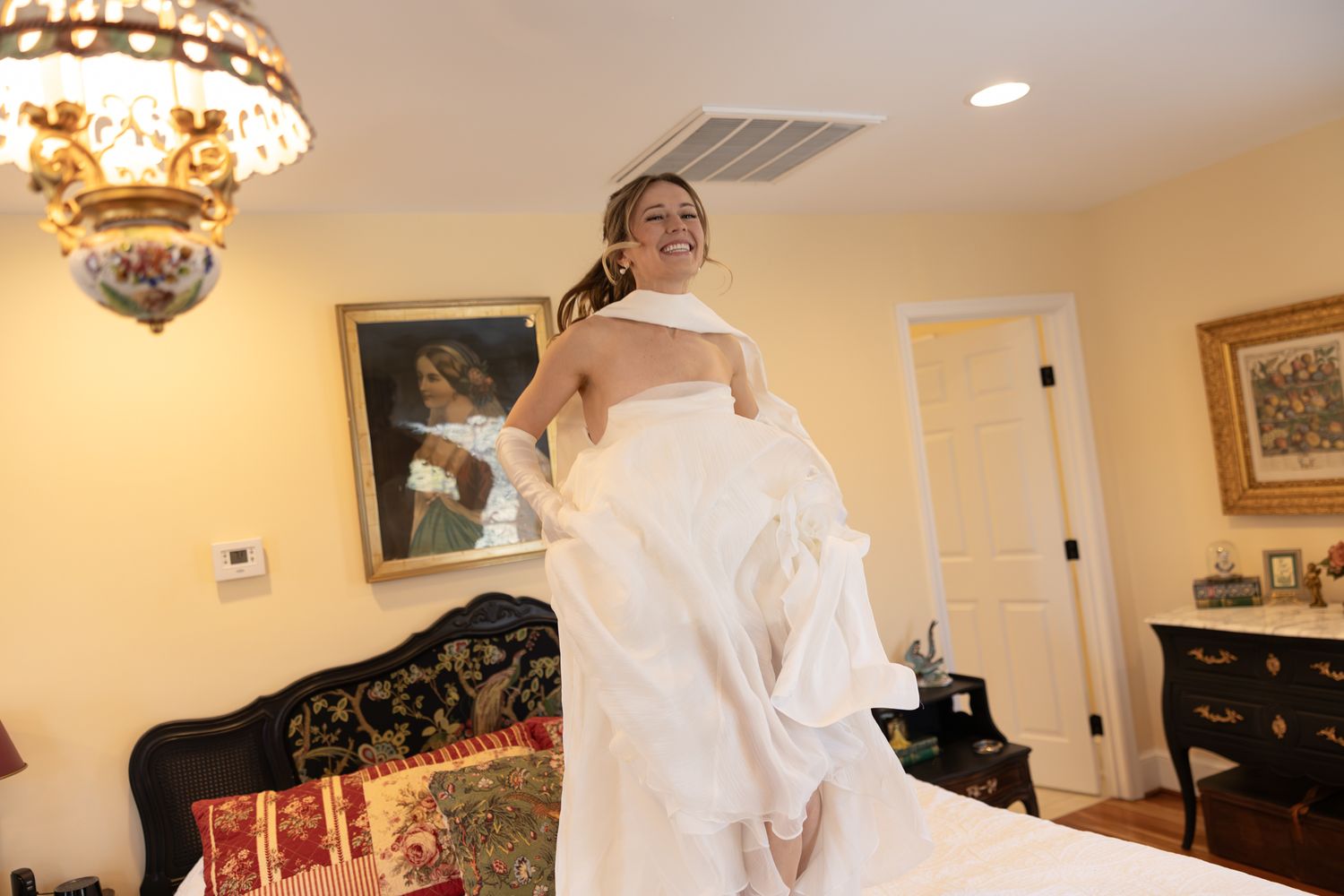 Smiling bride in white wedding gown lifting her dress in an elegantly decorated bedroom.