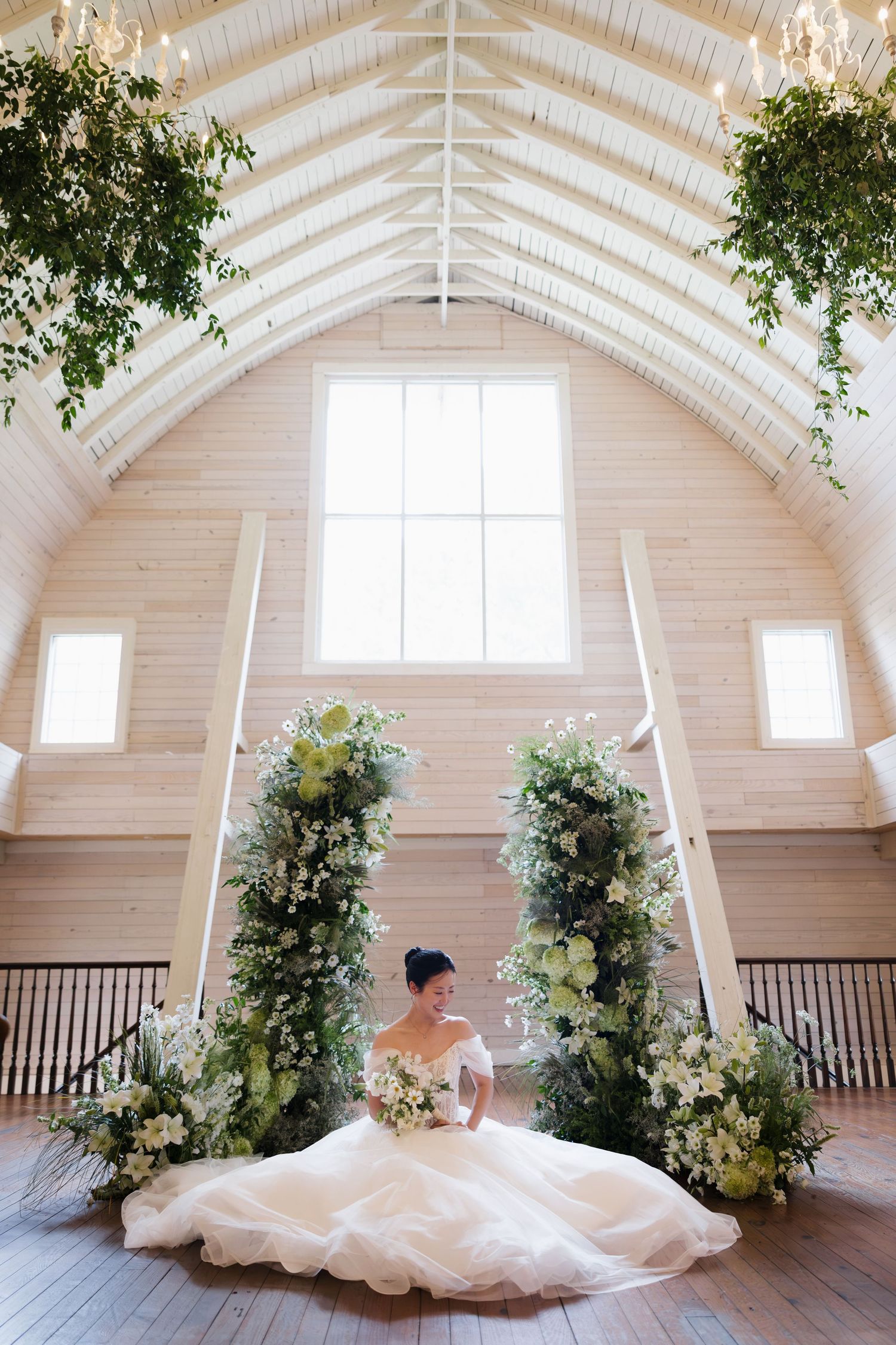Bride in a white ballgown sits before floral arch in a bright barn venue with vaulted ceilings and lush greenery.