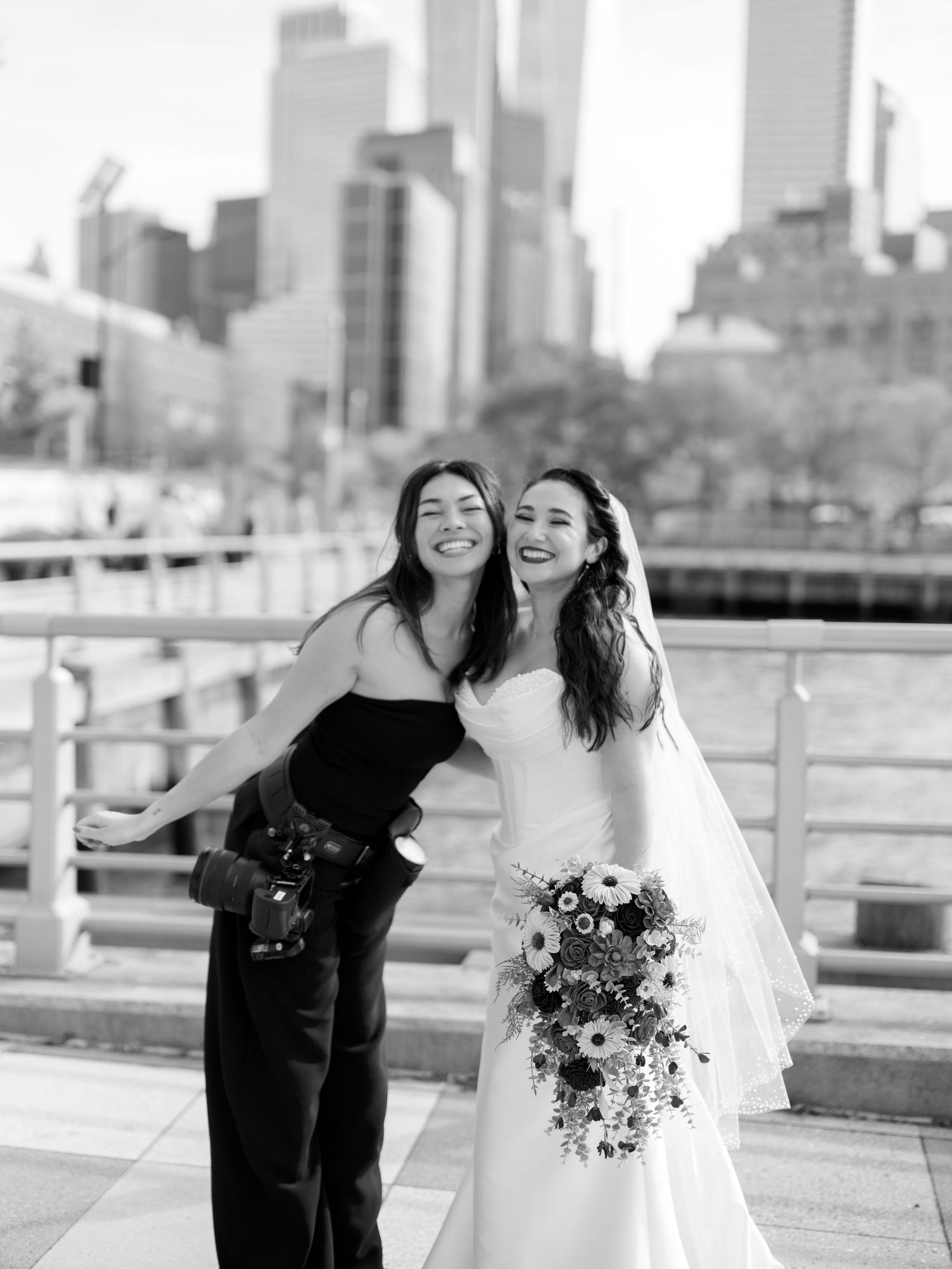 Black and white photo of a bride in a white gown holding a bouquet posing with a friend against NYC skyline.