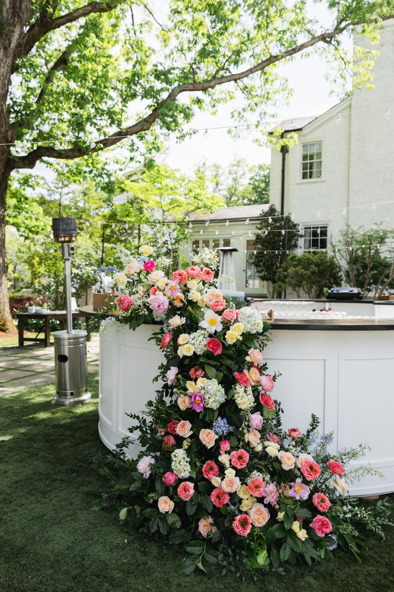 A lush cascading floral arrangement of pink and peach roses decorates a white outdoor bar at a garden wedding.