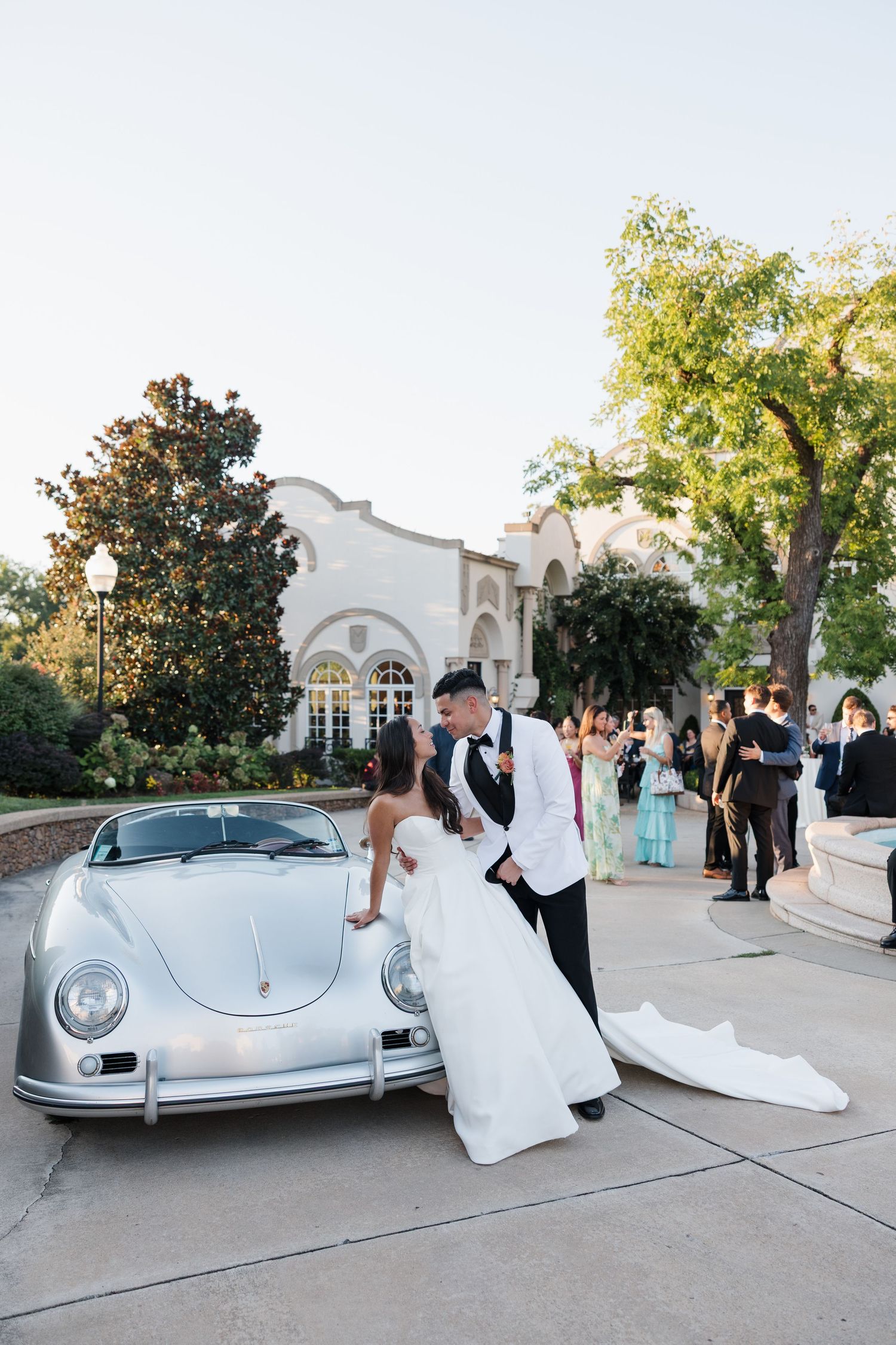 Bride and groom share a romantic moment beside a classic silver Porsche 356 convertible at an elegant outdoor wedding venue.