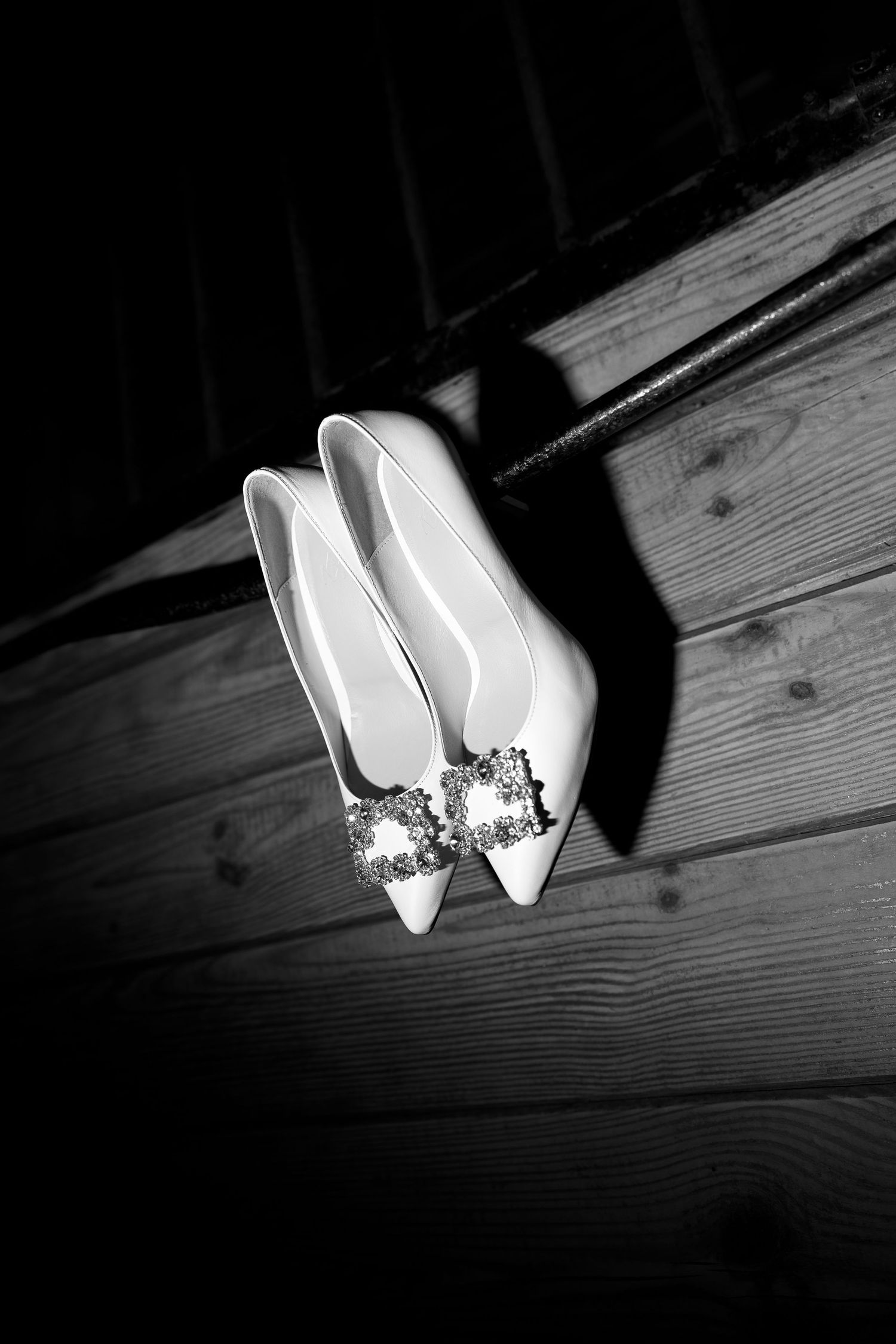 Black and white photo of elegant white bridal heels with crystal buckles displayed on rustic wooden steps.