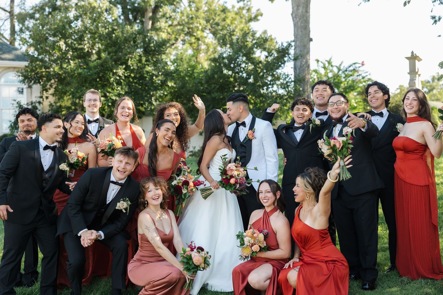 Wedding party poses outdoors, bride and groom surrounded by bridesmaids in red dresses and groomsmen in black tuxedos.