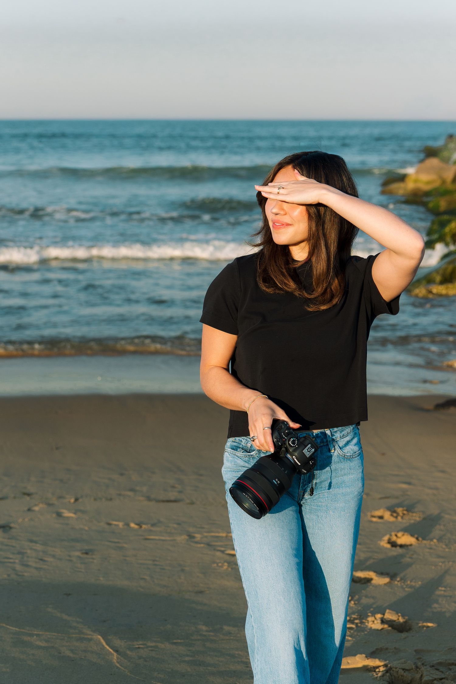 Young female photographer holding a DSLR camera on a sandy beach at sunset shielding her eyes from sun.