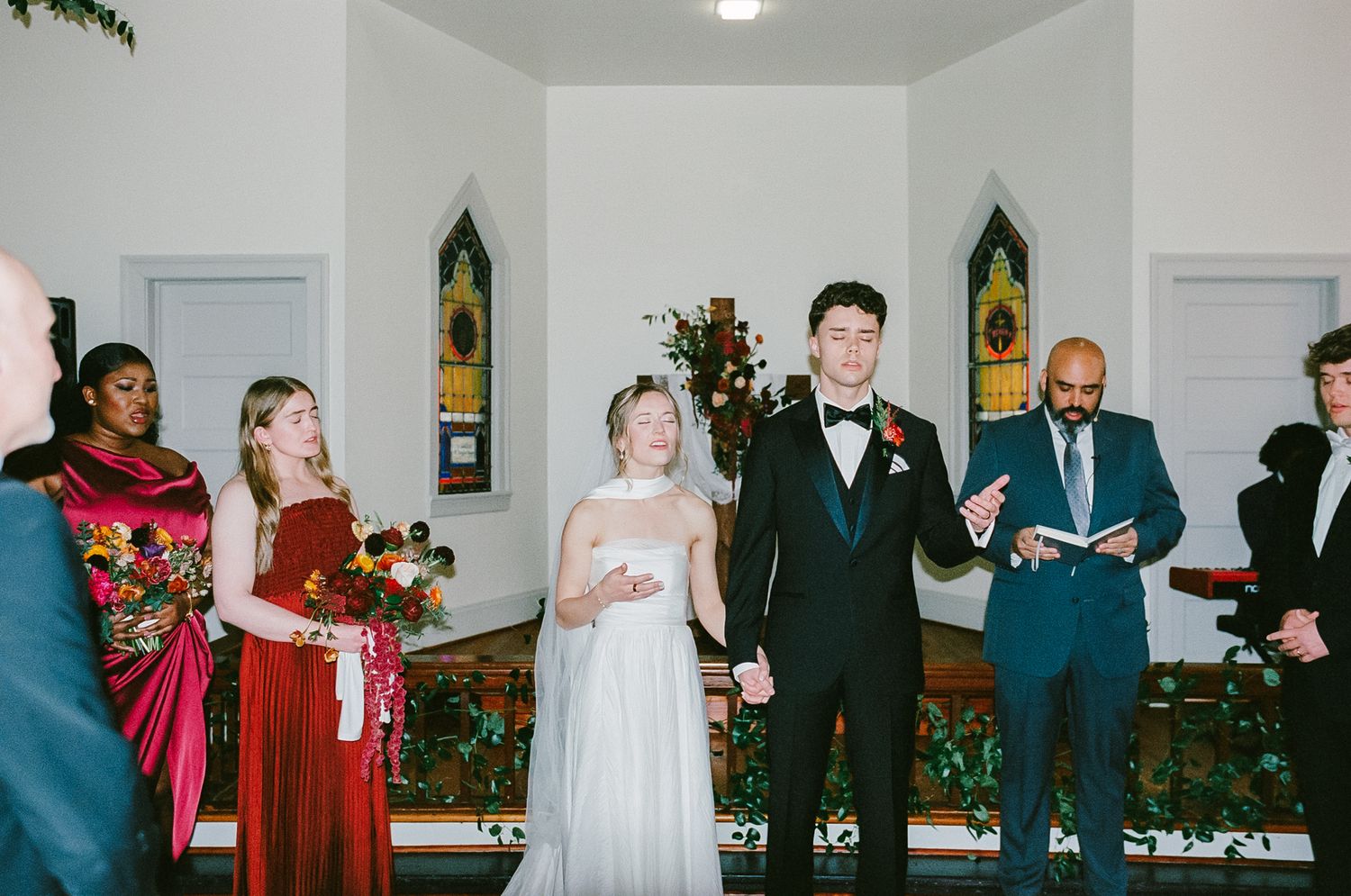 Bride and groom holding hands at wedding altar with bridesmaids in red dresses photographed on 35mm film.