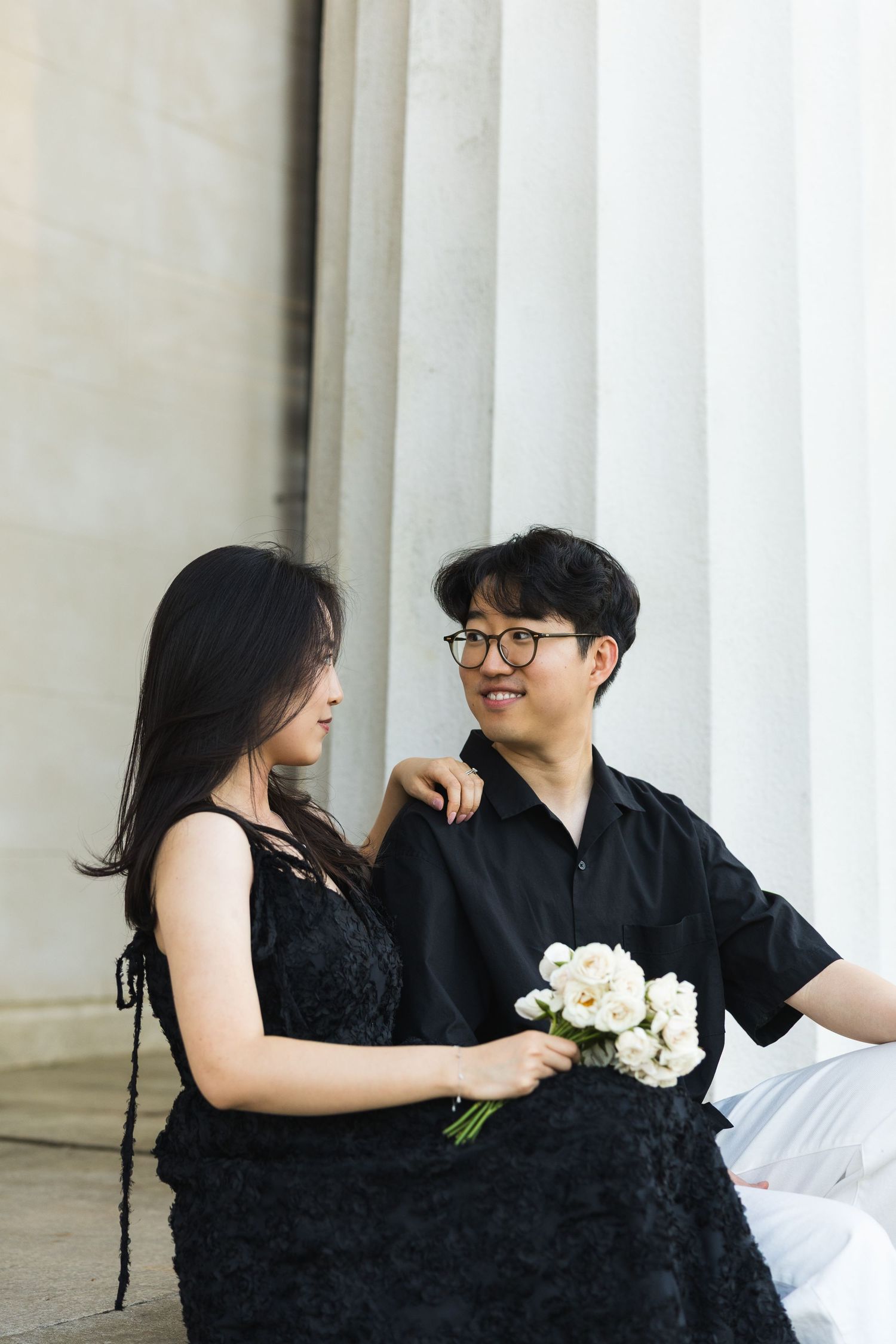 A smiling couple sits together on steps by white columns, woman holding white flower bouquet.