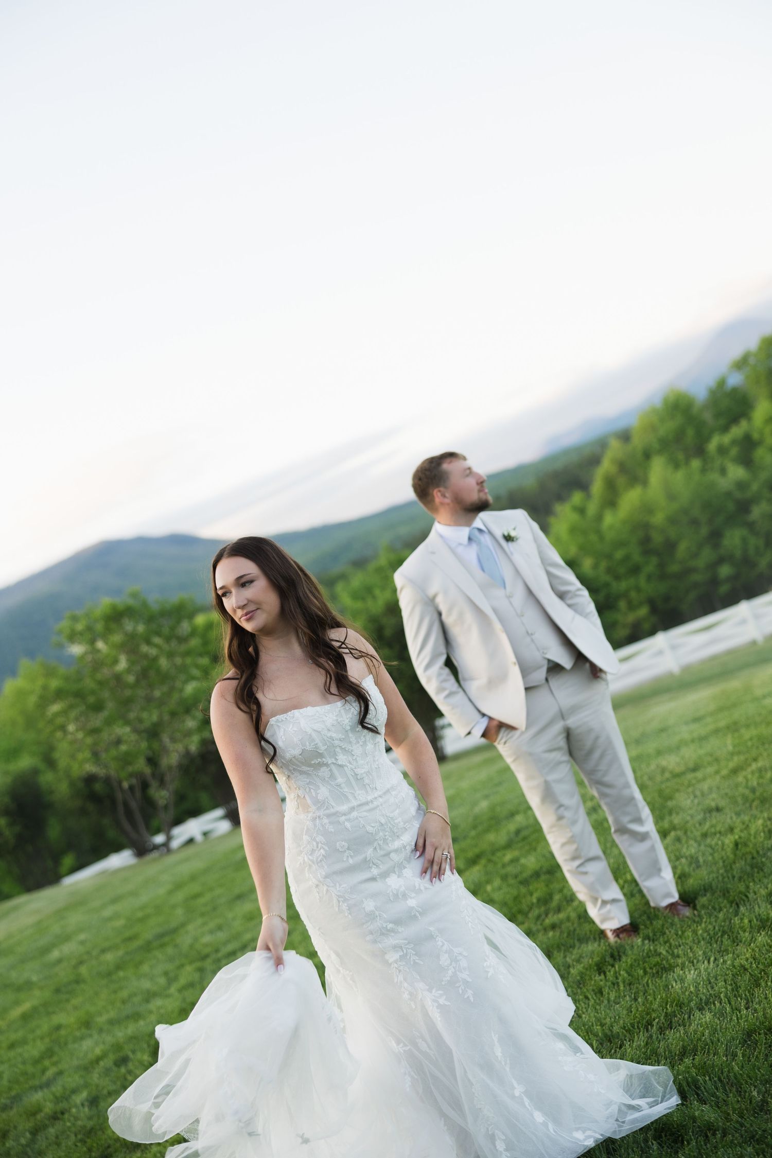 Bride in lace mermaid gown stands elegantly while groom looks away on green hillside.