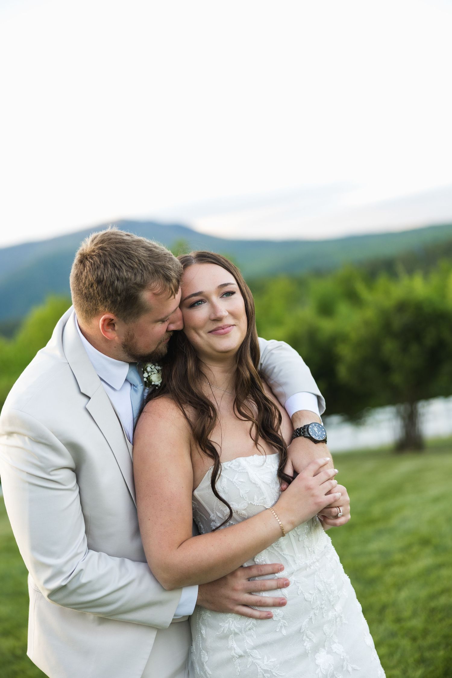 Groom in tan suit embraces bride in strapless lace gown with mountain backdrop at sunset.